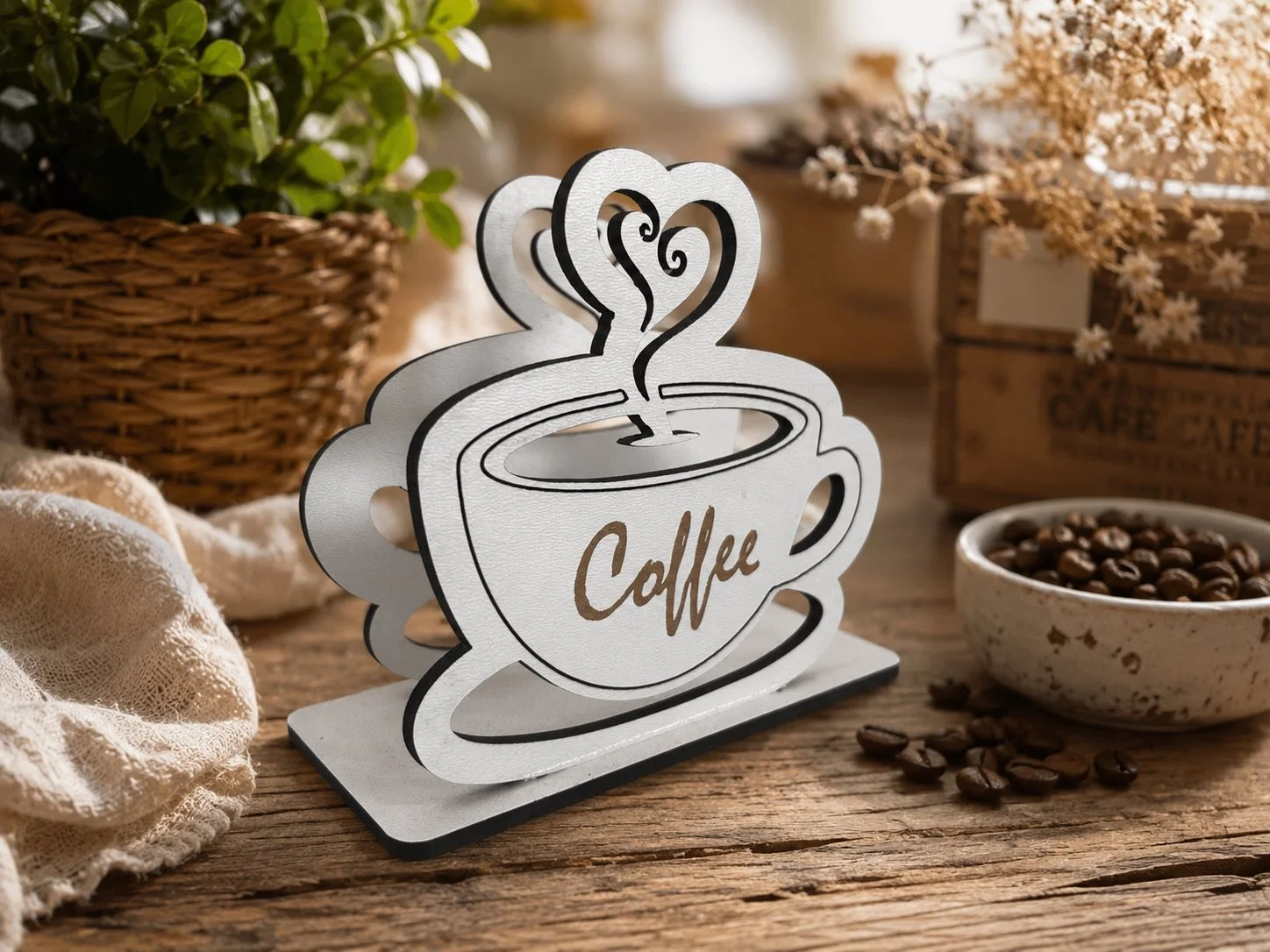 Decorative metal coffee cup with steam design and the word "Coffee" written on it, placed on a wooden surface surrounded by coffee beans, a bowl of coffee beans, a basket with green leaves, and a wooden box, with flowers in the background.