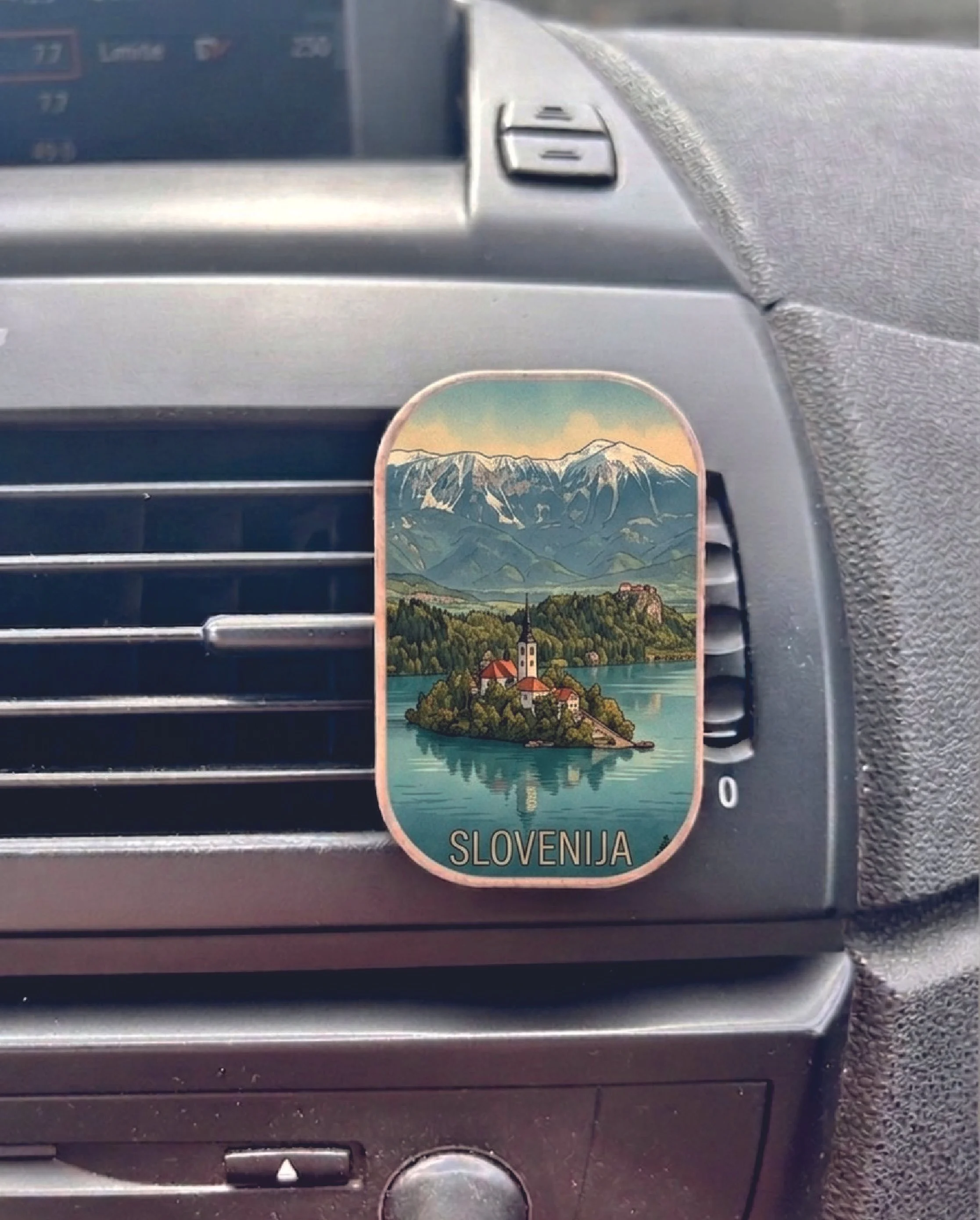 Slovenia-themed souvenir sticker on a car vent, depicting a mountain landscape with a church, lake, and village, with the label 'Slovenija.'