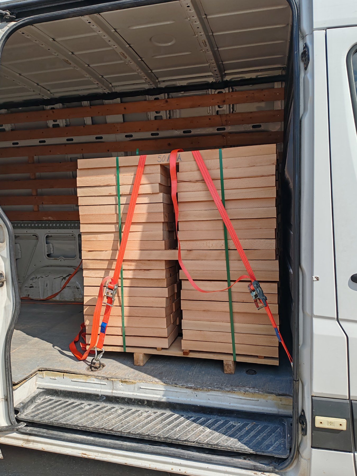 The back of a white cargo van filled with stacks of light-colored wooden planks secured with red and green straps.