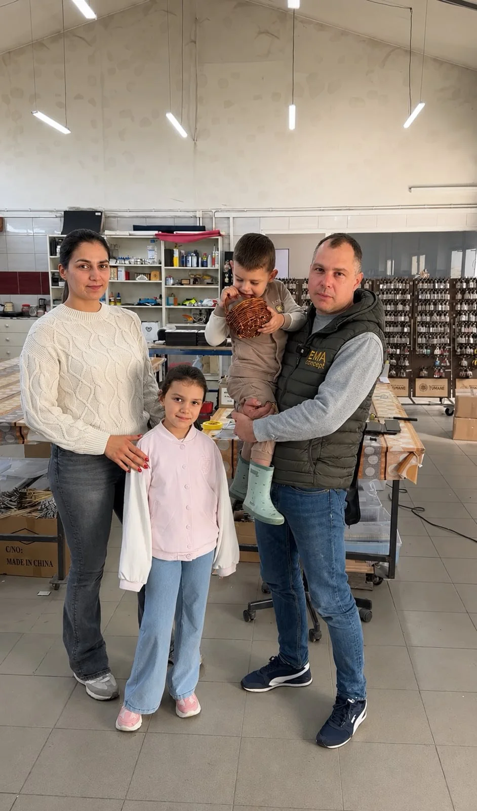 A family of four inside a workshop or manufacturing facility, with shelves and work tables in the background. The woman is standing next to a young girl, the man is holding a young boy, all posing for the photo.
