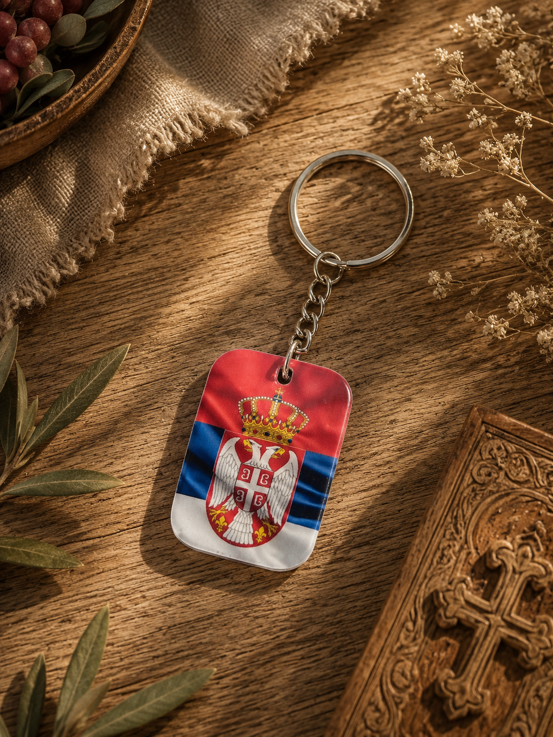 A metal keychain featuring the Serbian flag and coat of arms, resting on a wooden surface with decorative plants and a carved wooden box nearby.