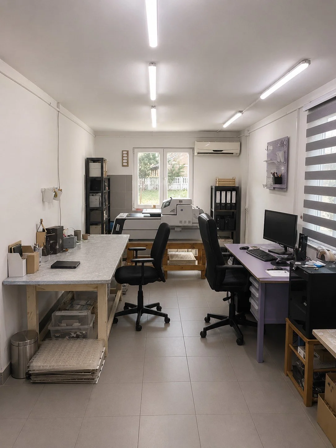 An organized office workspace with two desks, black office chairs, shelves with files, a computer, a printer, and office supplies, in front of a window with roller blinds and a view of a fenced yard.