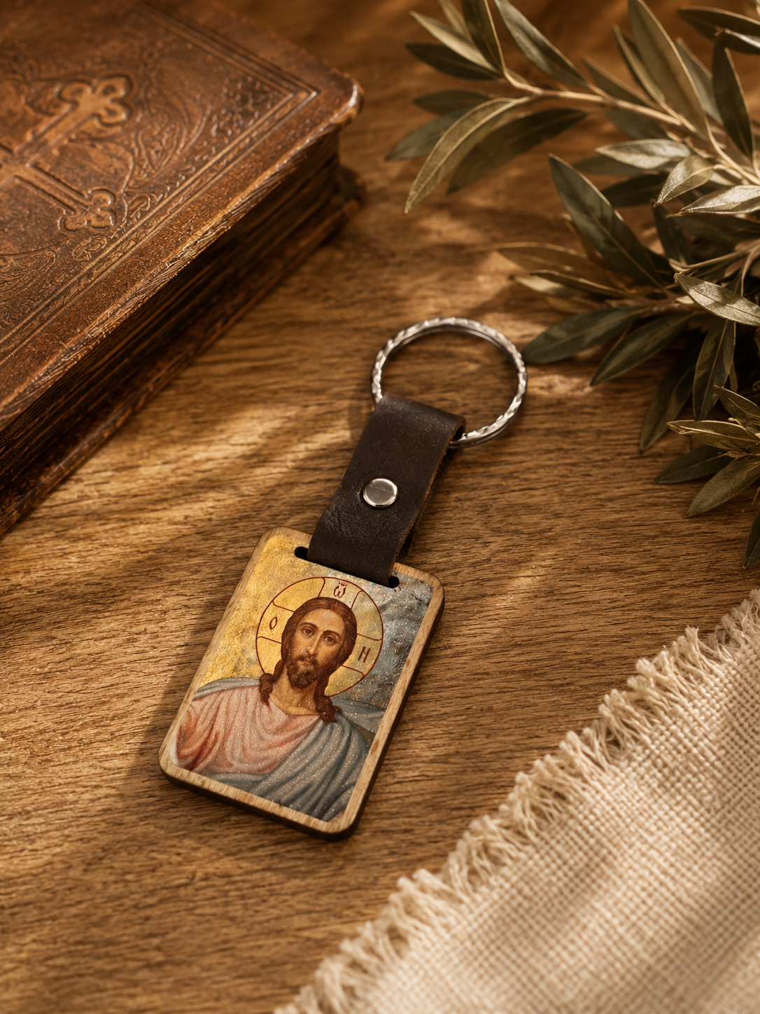 A religious pendant featuring an icon of Jesus Christ on a wooden keychain with a leather strap, placed on a wooden surface next to a book, some eucalyptus branches, and a piece of cloth.