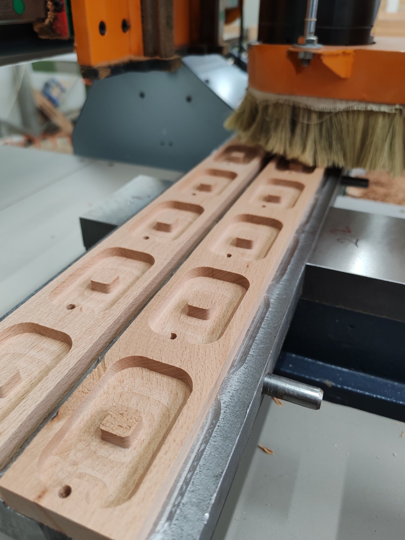 Close-up of a woodworking machine shaping a wooden piece with multiple rectangular and square cutouts, with a brush cleaning dust off the wood.