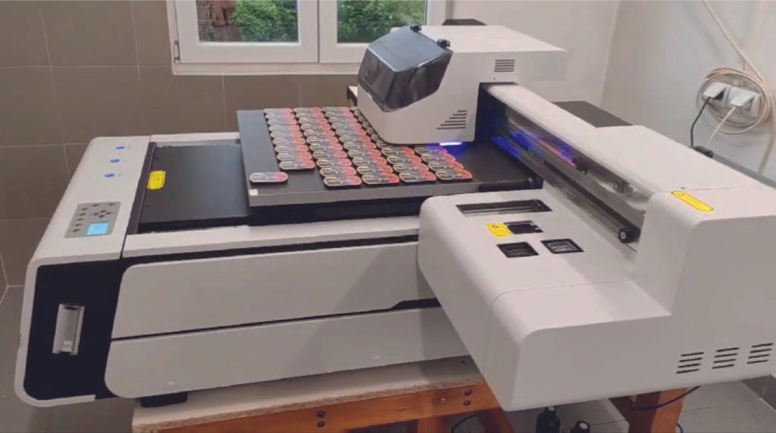 A high-tech machine for testing or sorting small electronic components, with numerous small circuit boards arranged in rows on the conveyor belt, situated in a room with a window showing greenery outside.