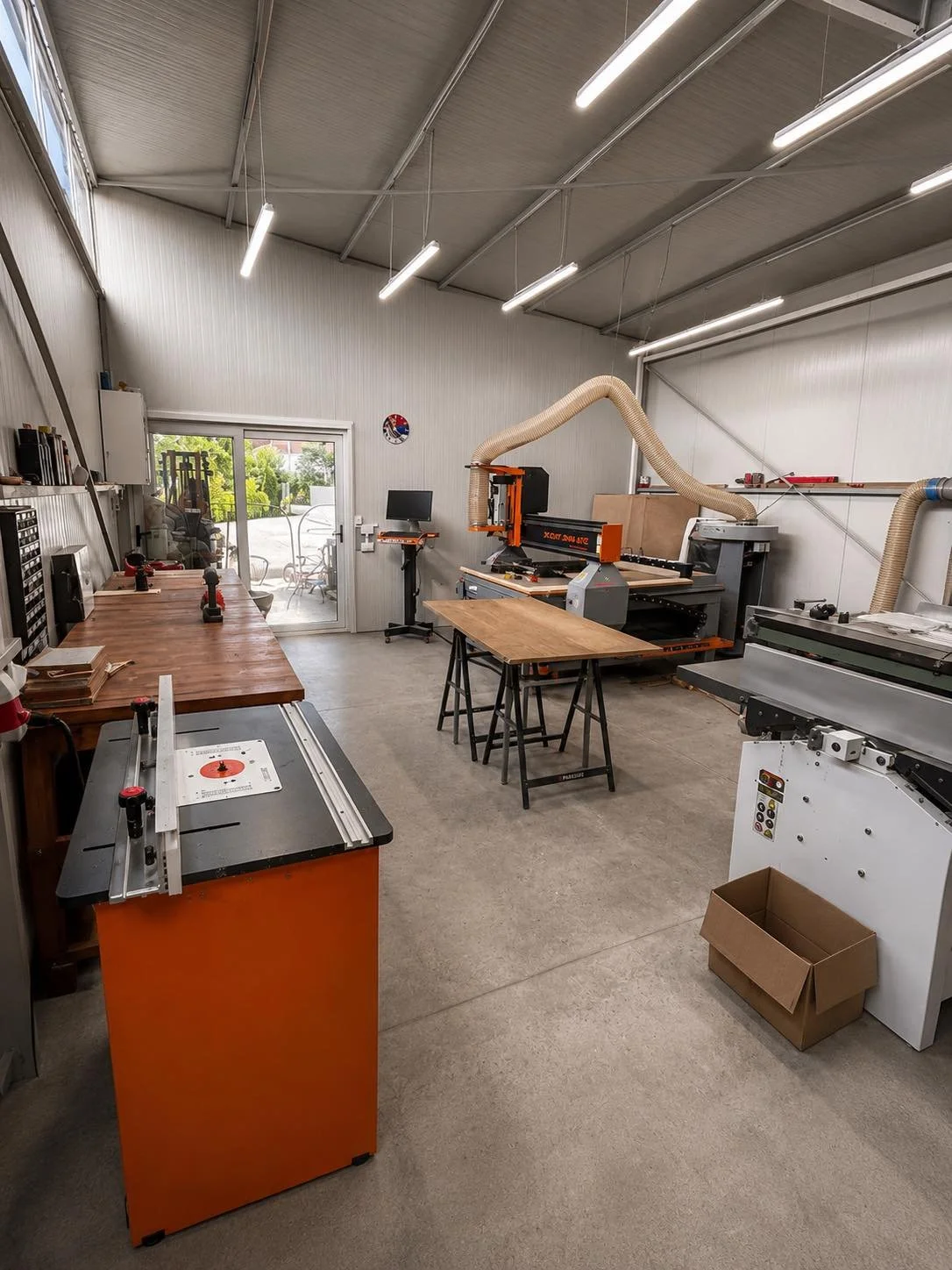 A woodworking workshop with various tools, a workbench, a router table, a CNC machine, and a sliding door leading outside.