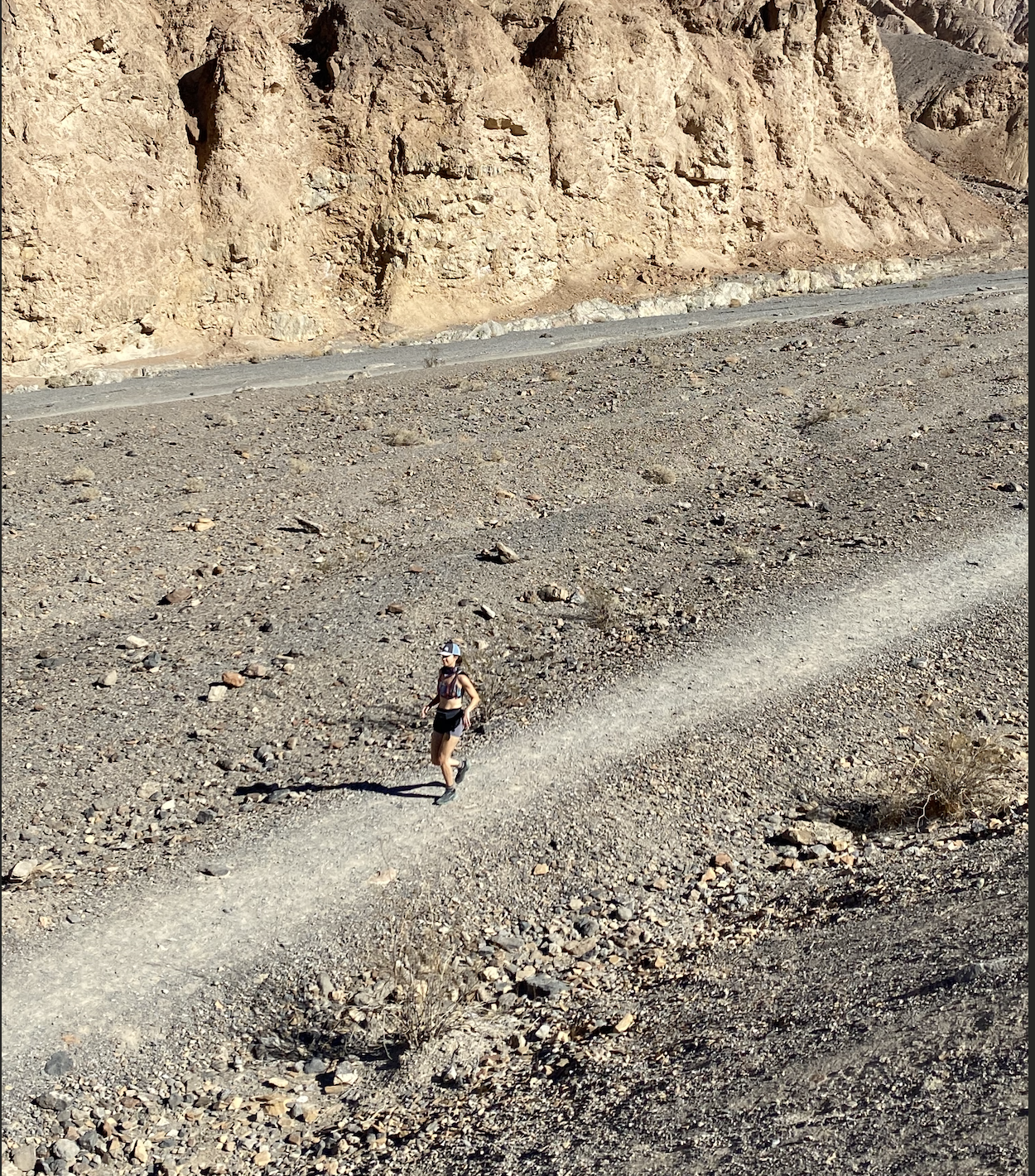 A woman endurance athlete wearing a hat, sunglasses, and athletic clothing is trail running on a dirt trail through a desert landscape in Death Valley with rocky terrain and large hills in the background.