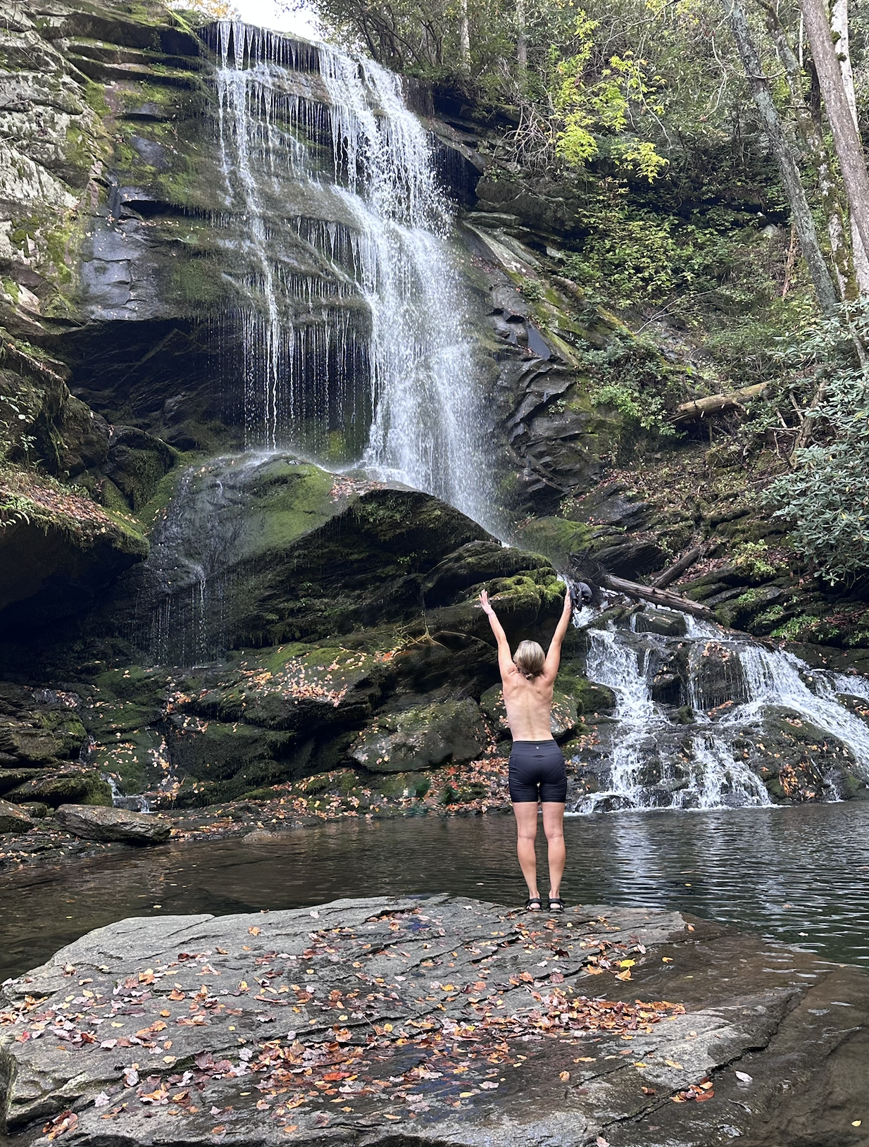 A person standing in a shallow pool of water with arms raised towards a waterfall in a forest setting. The waterfall cascades over rocks surrounded by green foliage during an excursion at a women's retreat.
