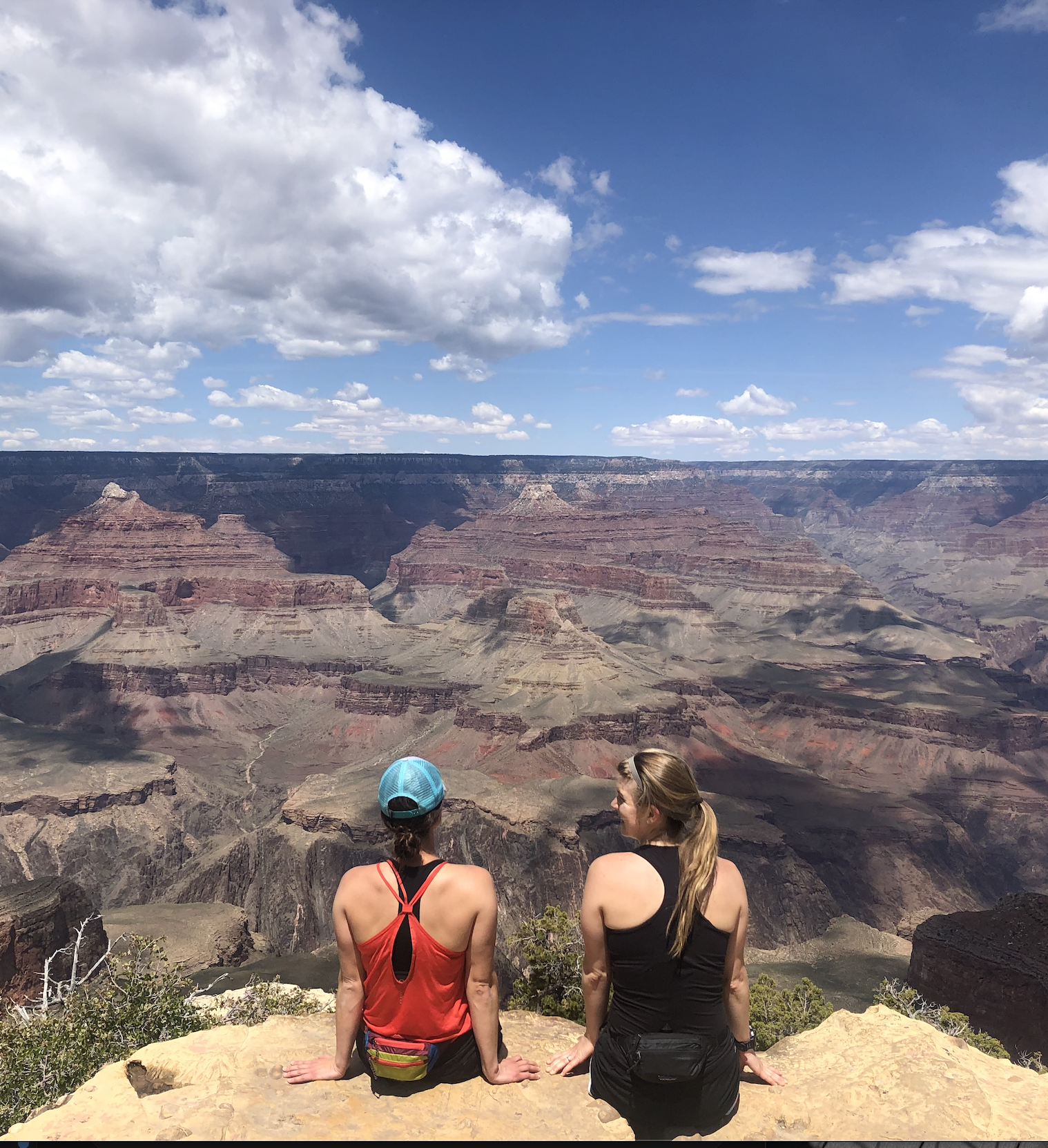 Two women sitting on a rock at the Grand Canyon, looking out over the vast canyon with layered rock formations under a partly cloudy sky.