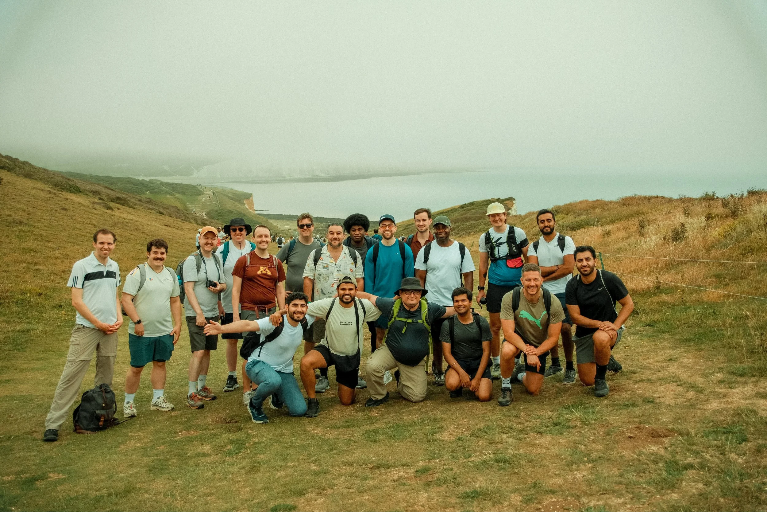 Group of hikers standing on a grassy hillside with coastline and cliffs in the background, some smiling and posing for the camera.