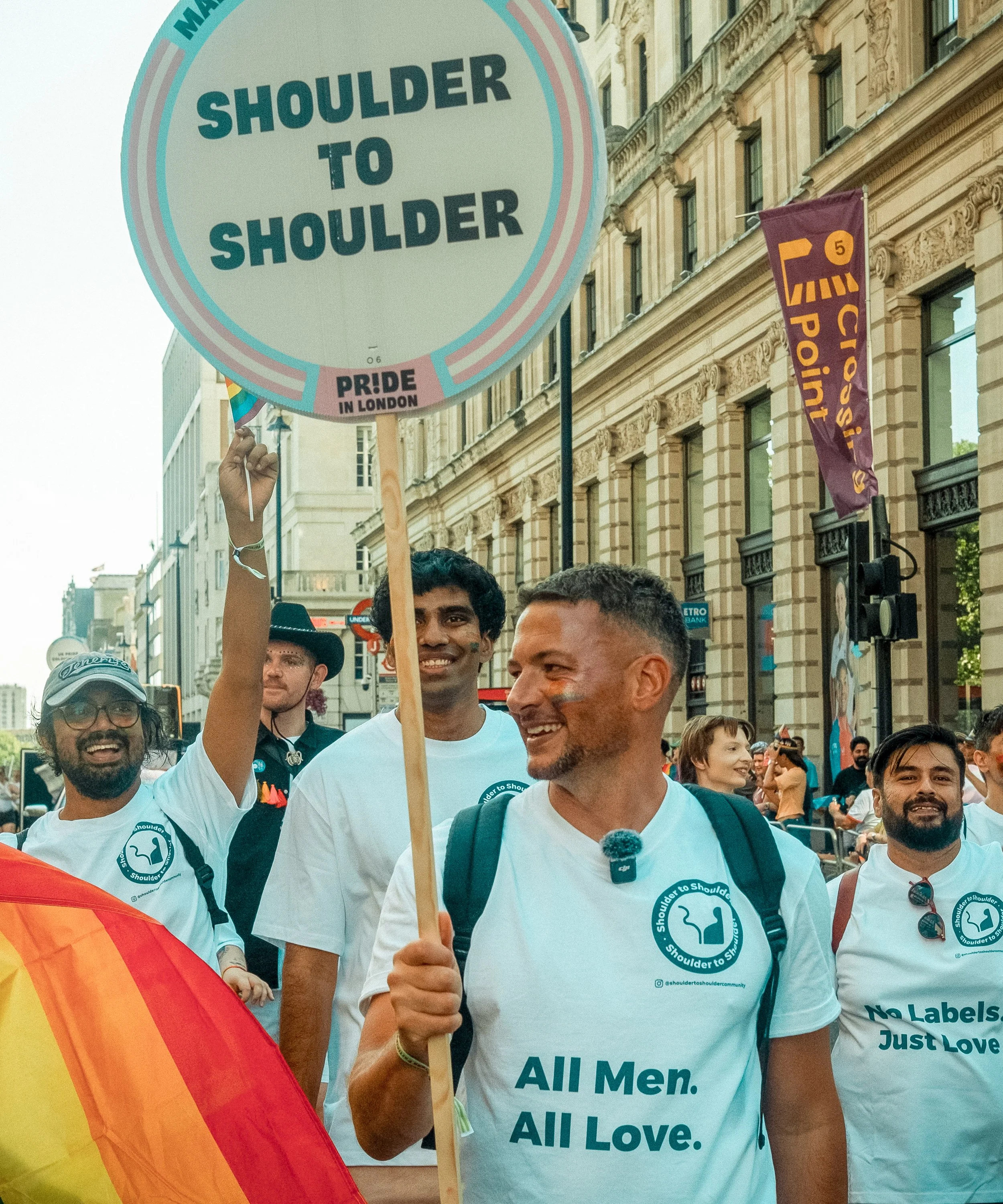 A group of diverse people participating in a pride parade, holding a sign that reads 'Shoulder to Shoulder,' and wearing white t-shirts with similar slogans. They celebrate LGBTQ+ pride in a city street with historic buildings and flags, including a rainbow flag. The atmosphere is joyful and inclusive.