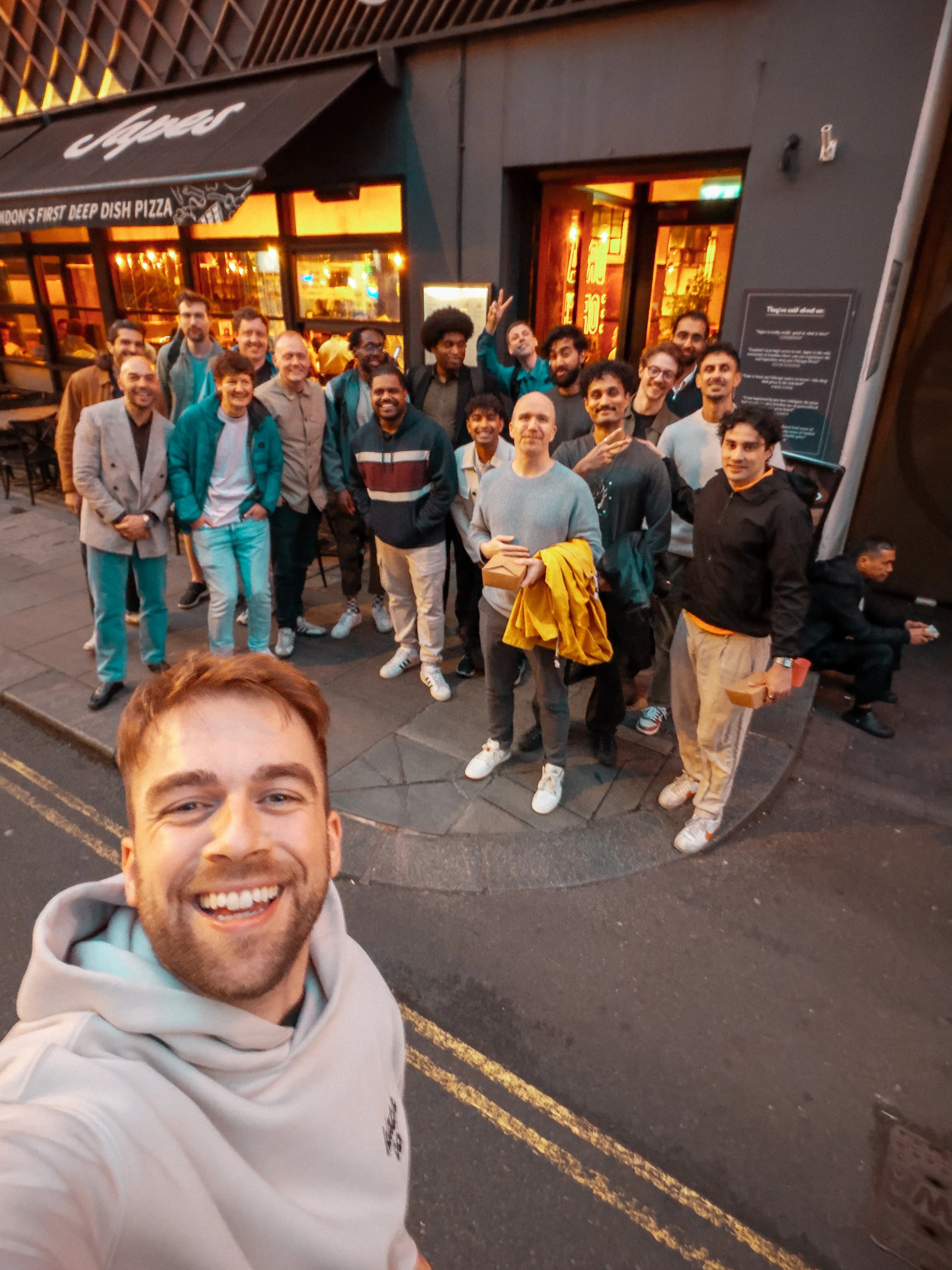 A group of people standing outside a restaurant on a city street at night, smiling and posing for a photo with a young man taking a selfie in the foreground.