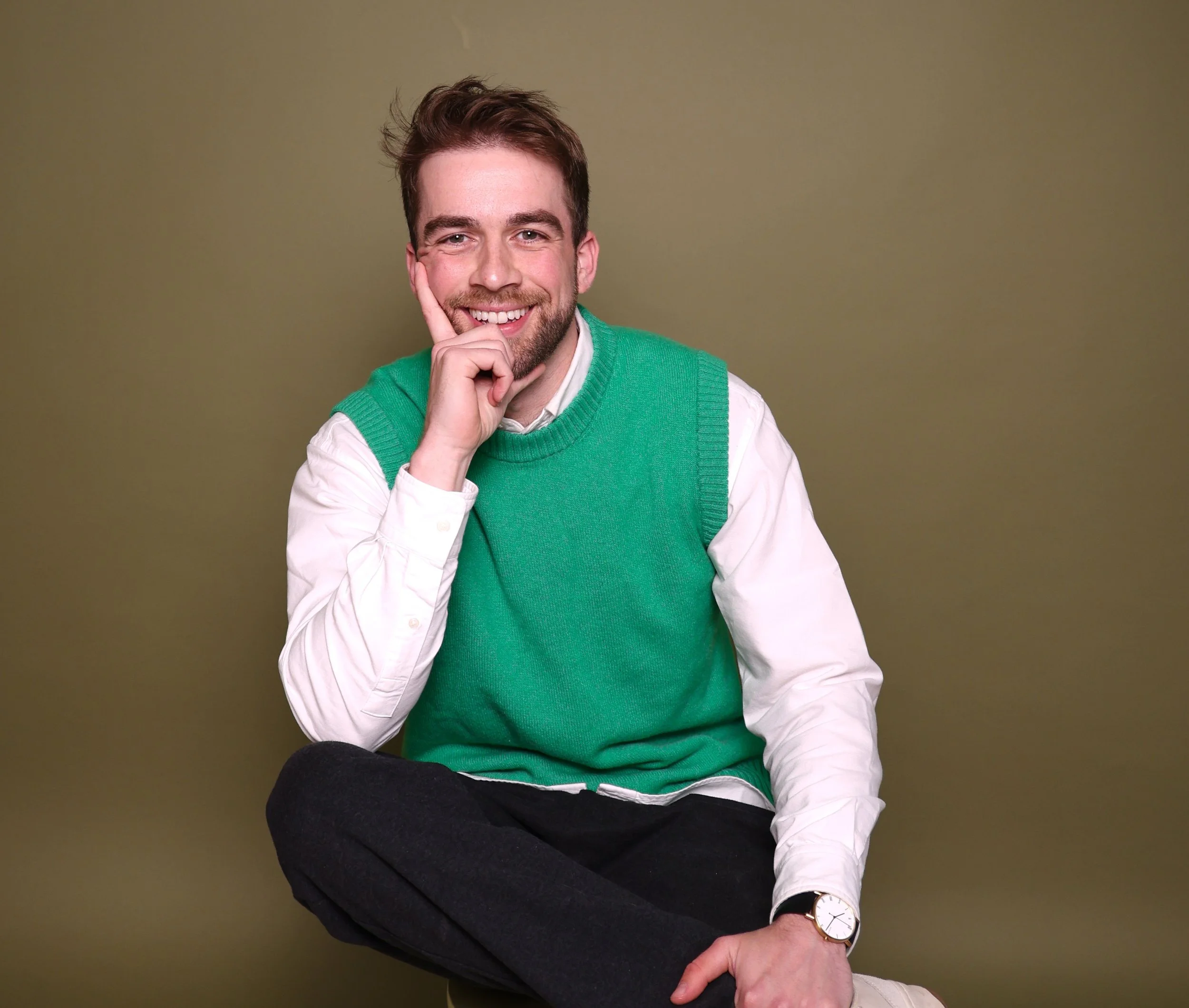 A smiling man with brown hair and a beard wearing a green vest over a white shirt, sitting against a plain greenish-beige background, with his hand resting on his cheek and his legs crossed.