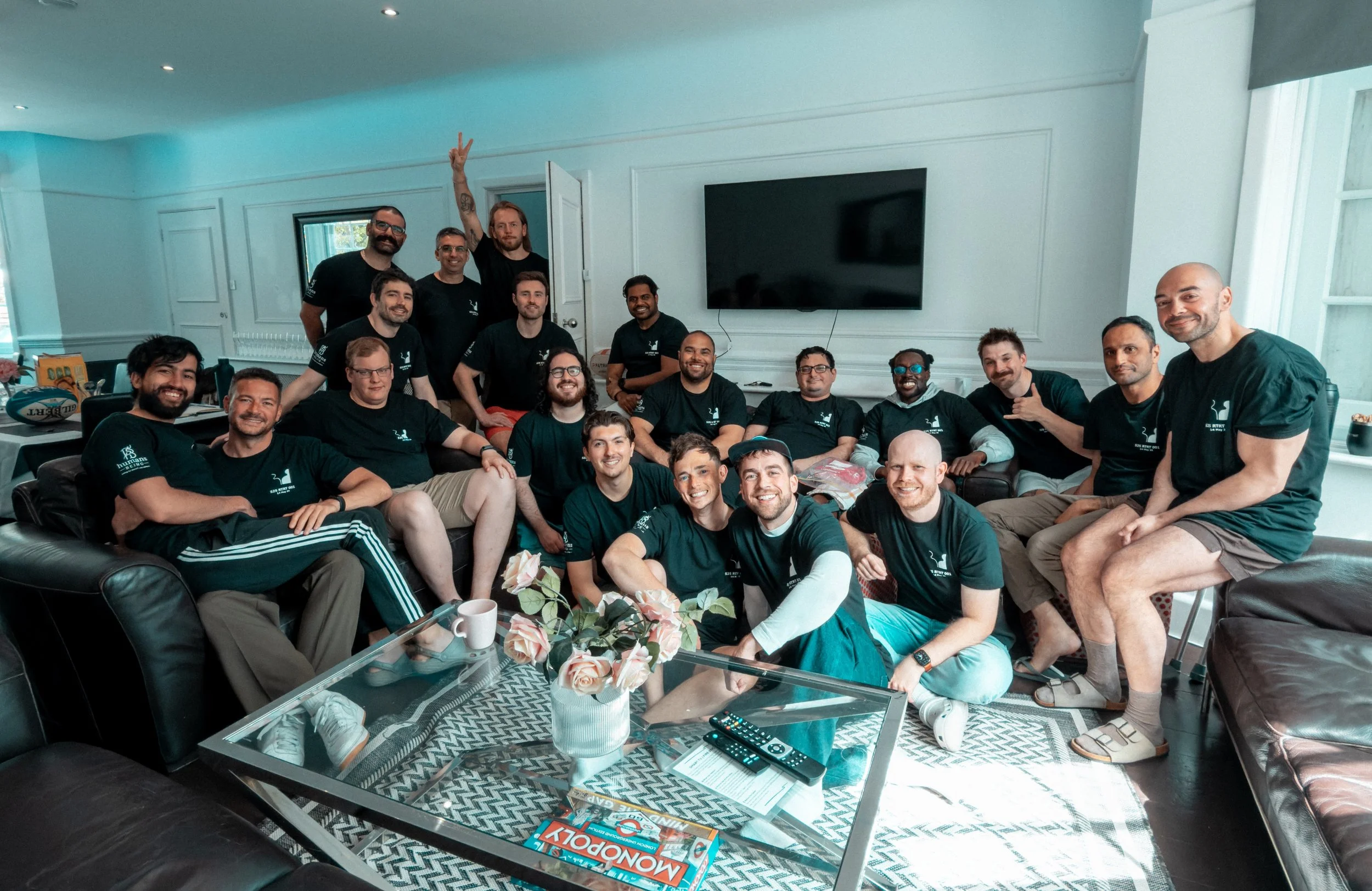 A group of 16 people in matching black t-shirts gathered in a living room for a photo. Some are seated on couches, others are standing behind them, and one person is making a peace sign. The room has a large flat-screen TV on the wall, and a glass coffee table with magazines, a flower arrangement, and remote controls in front of the group.