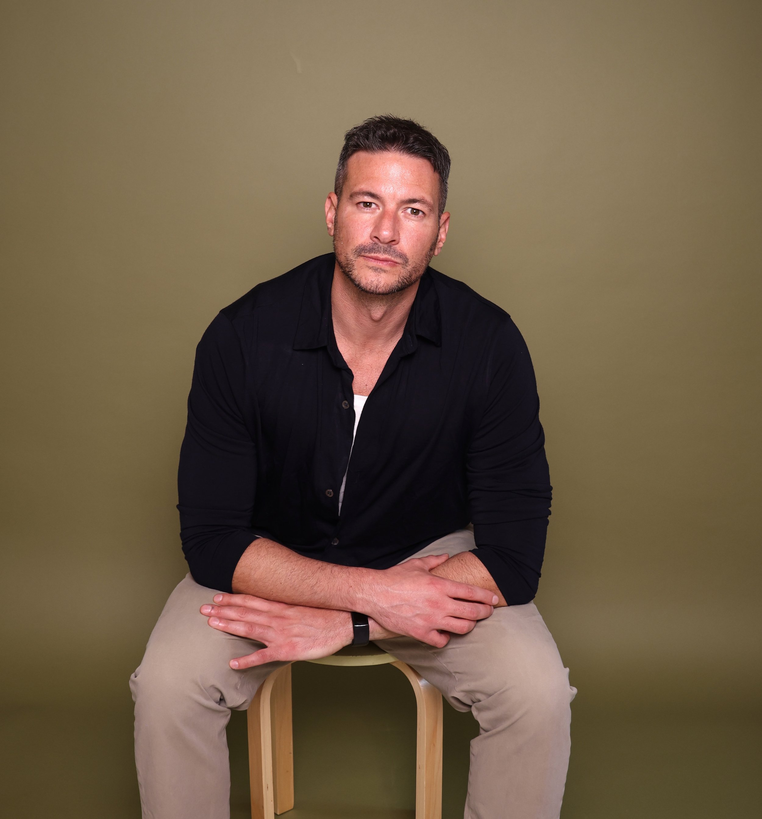 A man dressed in a black shirt and khaki pants sitting on a wooden stool, head tilted slightly, with a neutral expression, against an olive-green background.