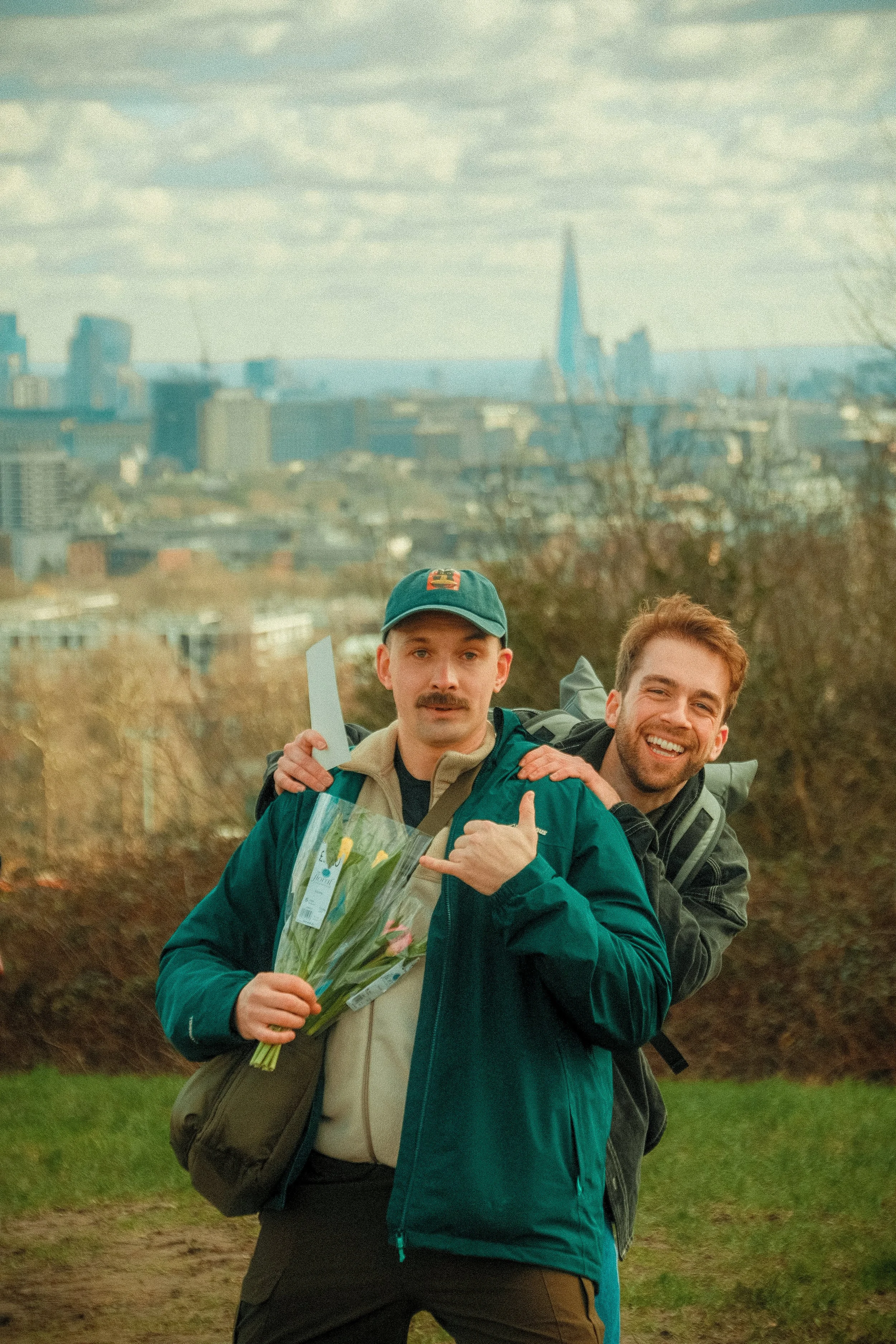 Two men walking outdoors, holding a bouquet of flowers, with a city skyline in the background.