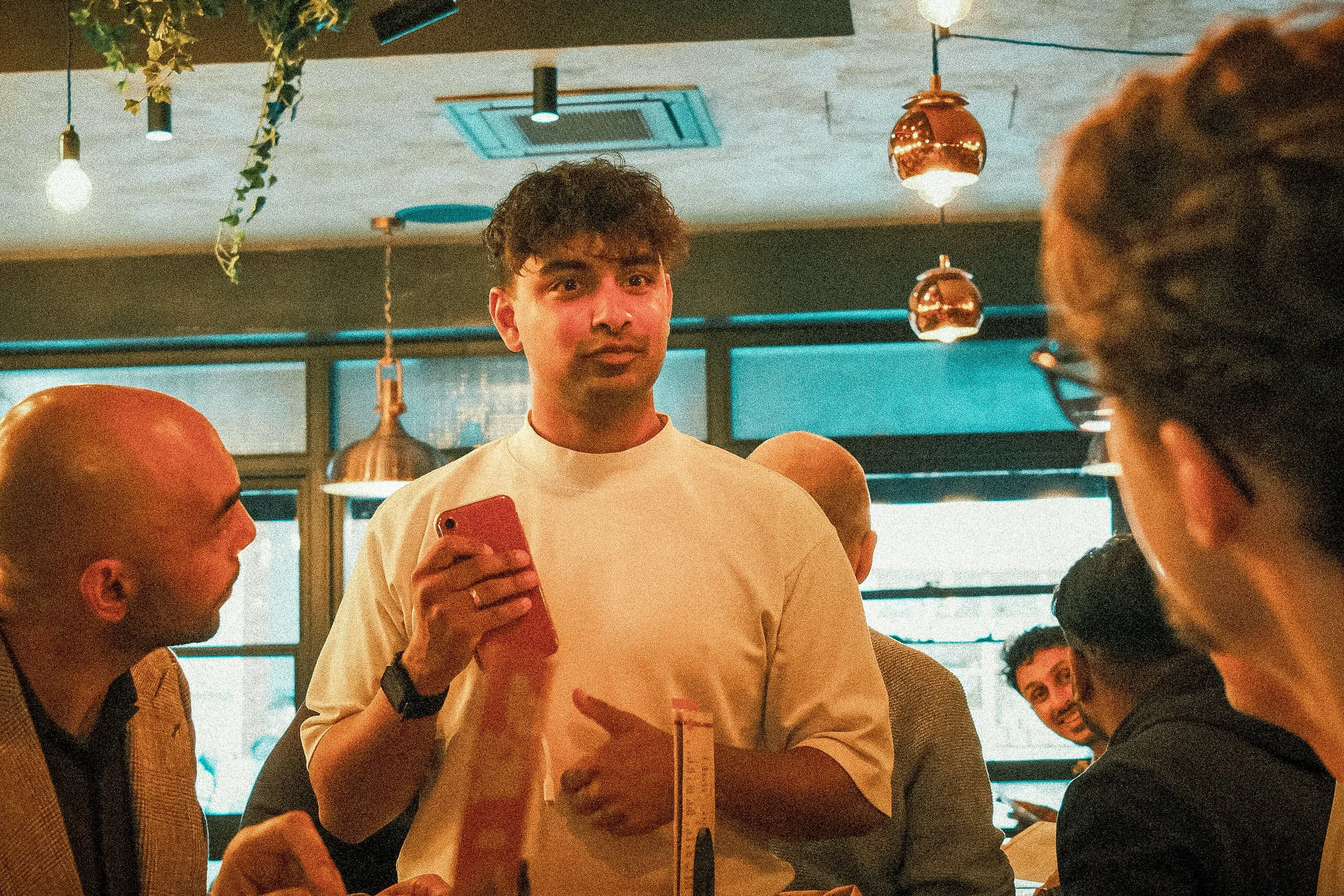 A young man with curly hair holding a red smartphone, talking to a group of people in a dimly lit modern restaurant or cafe, with hanging copper light fixtures and green plants.