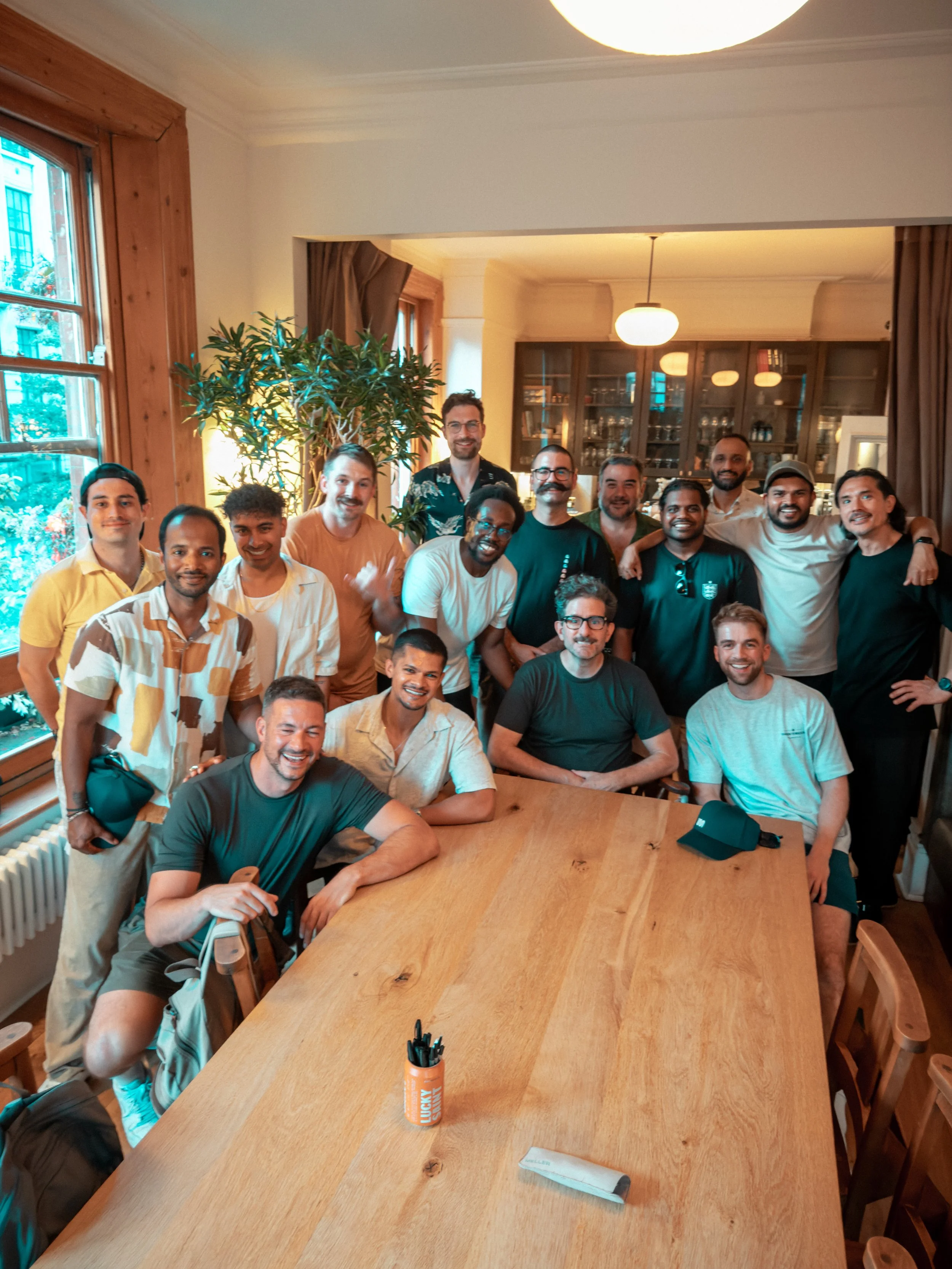 A group of 16 men gathered together in a cozy room with wood accents, smiling and posing for a photo. They are standing and sitting around a large wooden table, with some wearing casual shirts and others in T-shirts. There are two visible hats on the table and a cup of pens. The room has large windows with curtains and a glass cabinet filled with glasses and bottles in the background.