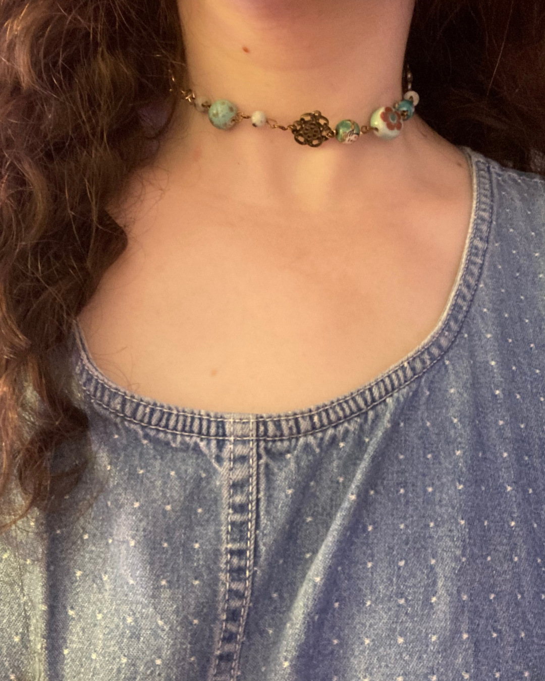 Close-up of a person wearing a beaded necklace with a decorative pendant and a denim shirt with a rounded neckline.