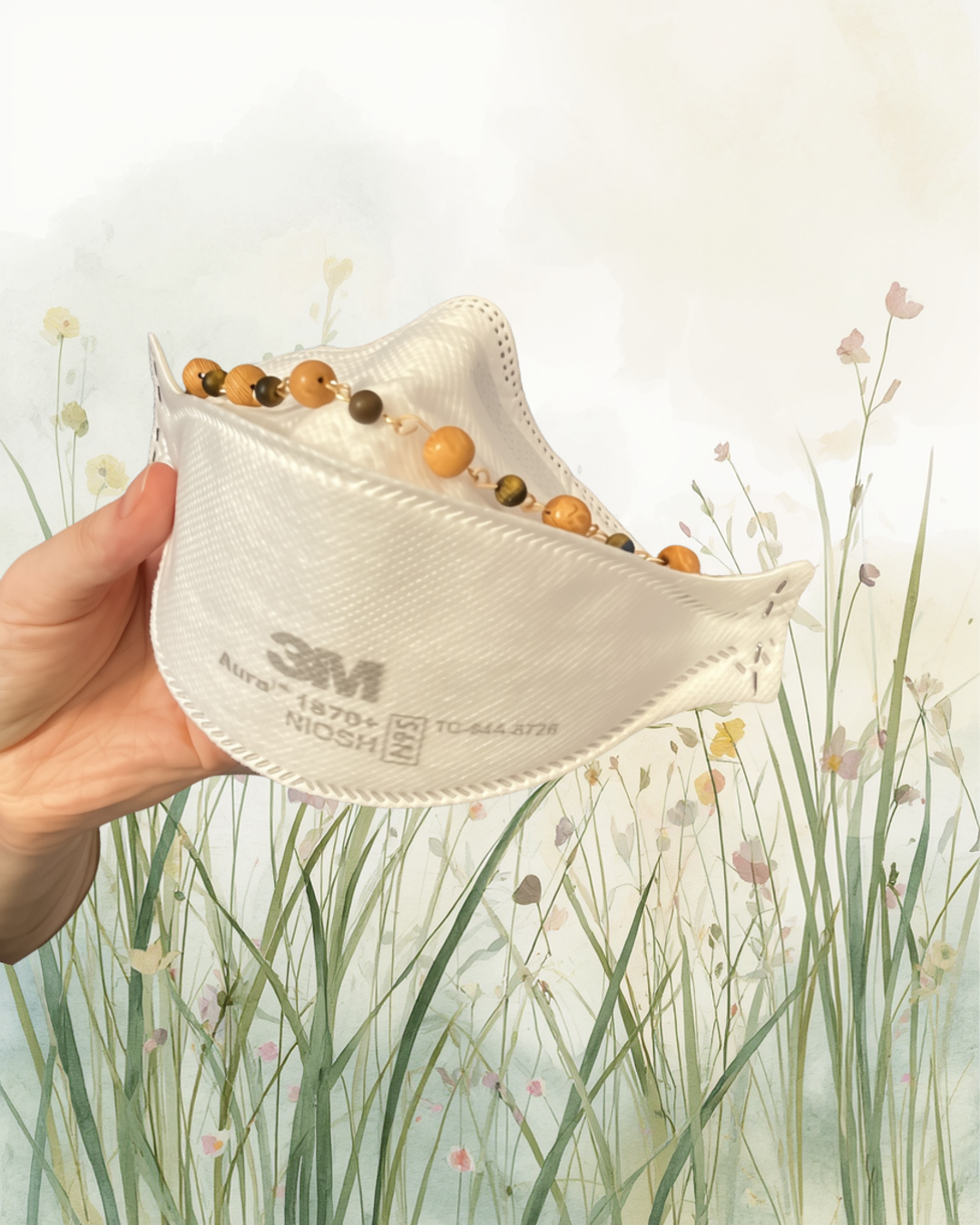 Person holding a white 3M 1870-N95 respirator mask with a beaded necklace resting inside it, against a background with a pastel-colored floral and grass pattern.