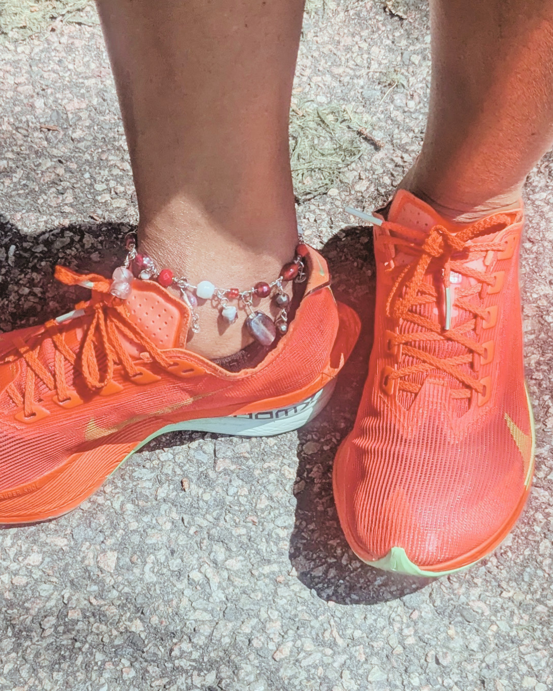 A person's feet wearing orange running shoes and an anklet with beads, standing on a gravel surface. Can also be worn as a mask chain