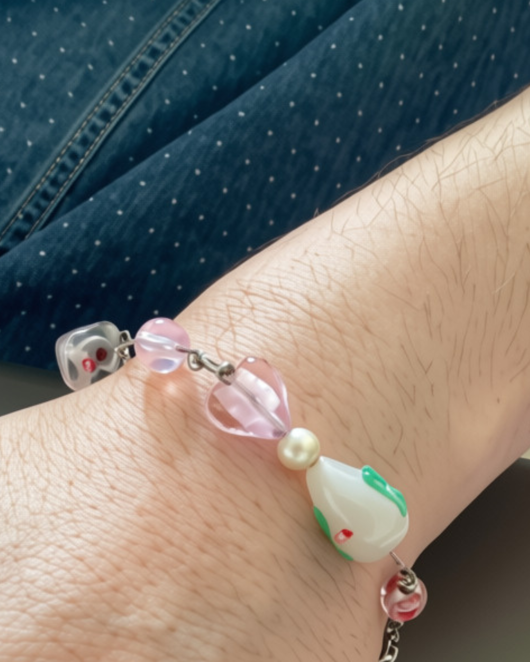 A wrist with a bracelet made of various beads, including pink, white, pearl, and decorative ones, against a background of a blue denim fabric.
