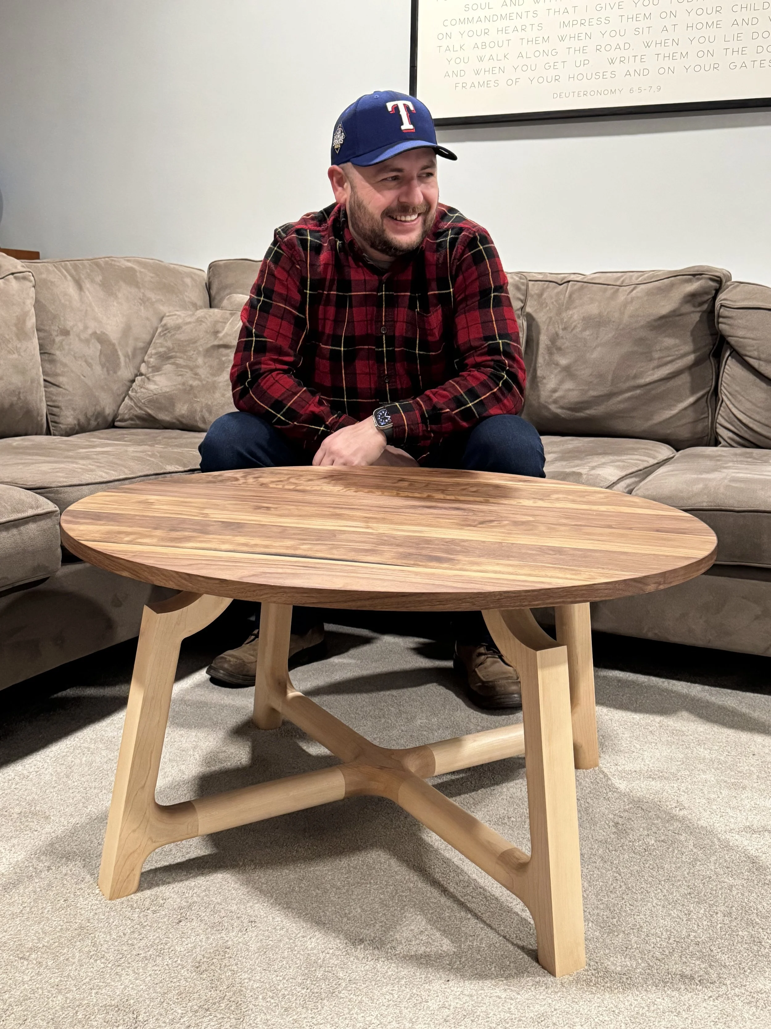 A smiling man sitting on a beige sofa, wearing a red and black plaid shirt, jeans, brown shoes, a blue baseball cap with a red T, and a watch, in a living room with a wooden table and a framed print on the wall.