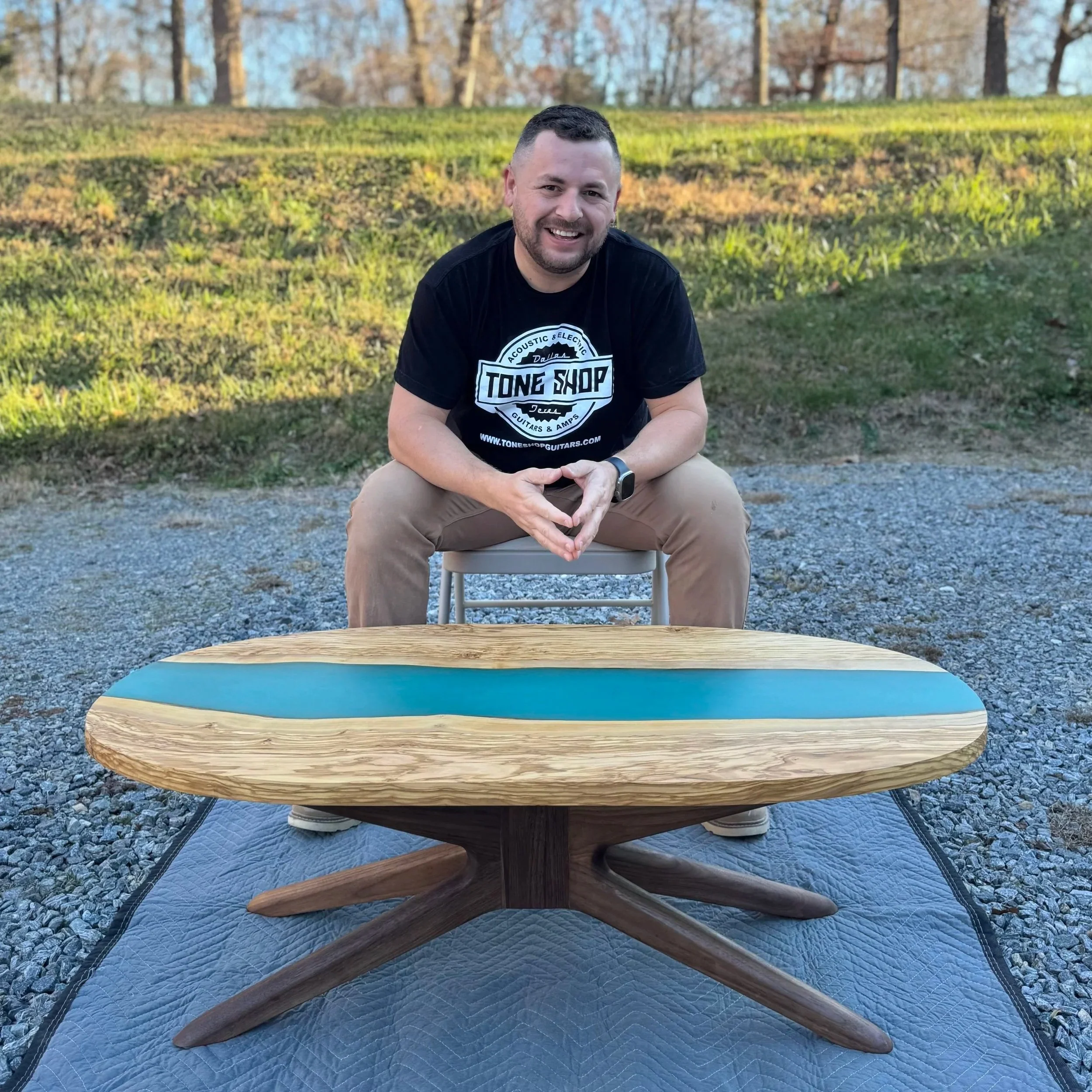 A smiling man with short dark hair and a beard, wearing a black T-shirt with a logo, is sitting outside on a chair. In front of him, there is a wooden table. The background shows a grassy area with trees, indicating a park or backyard setting during late afternoon or early evening.