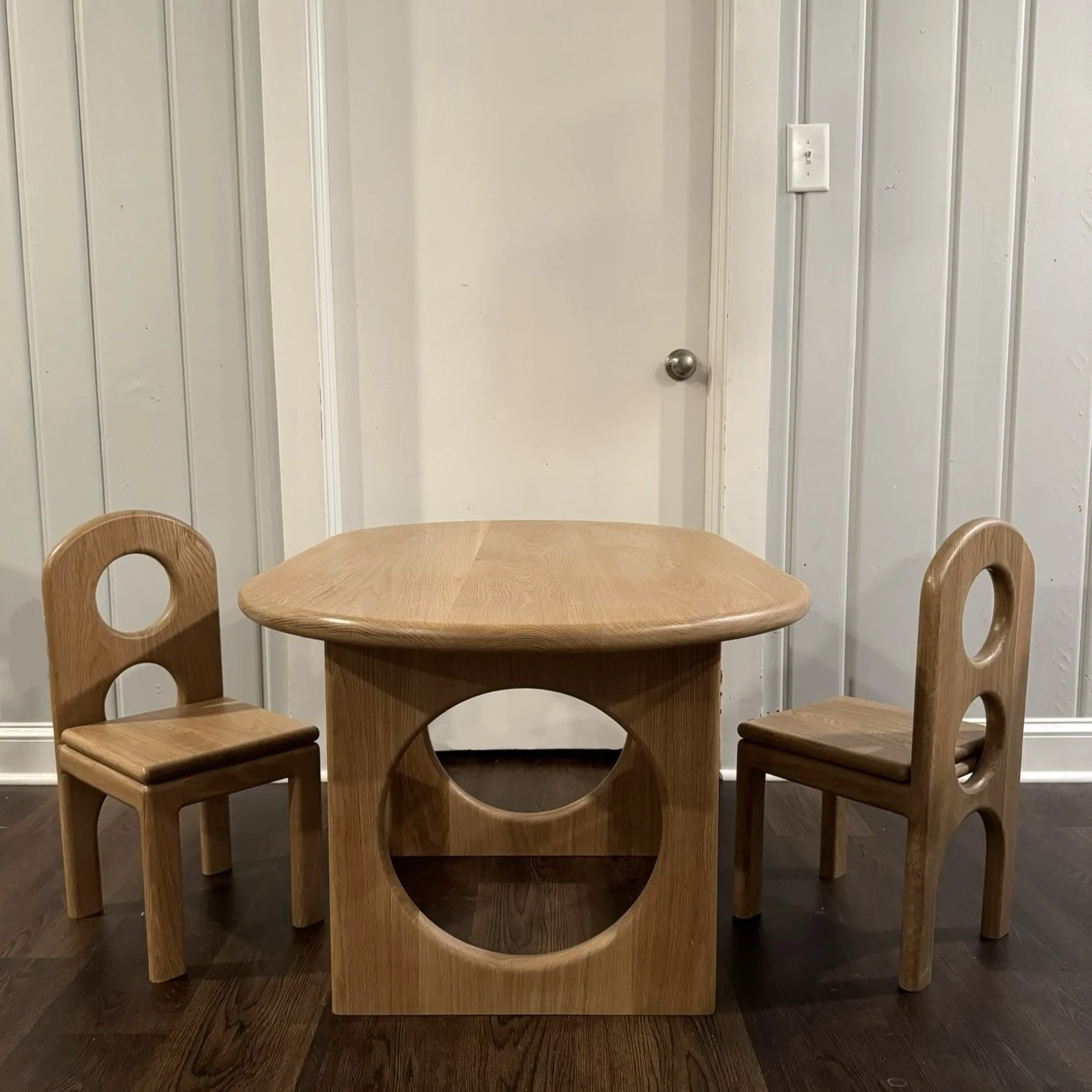 A kids wooden dining table with two matching chairs in a room with white paneled walls and dark hardwood floor.