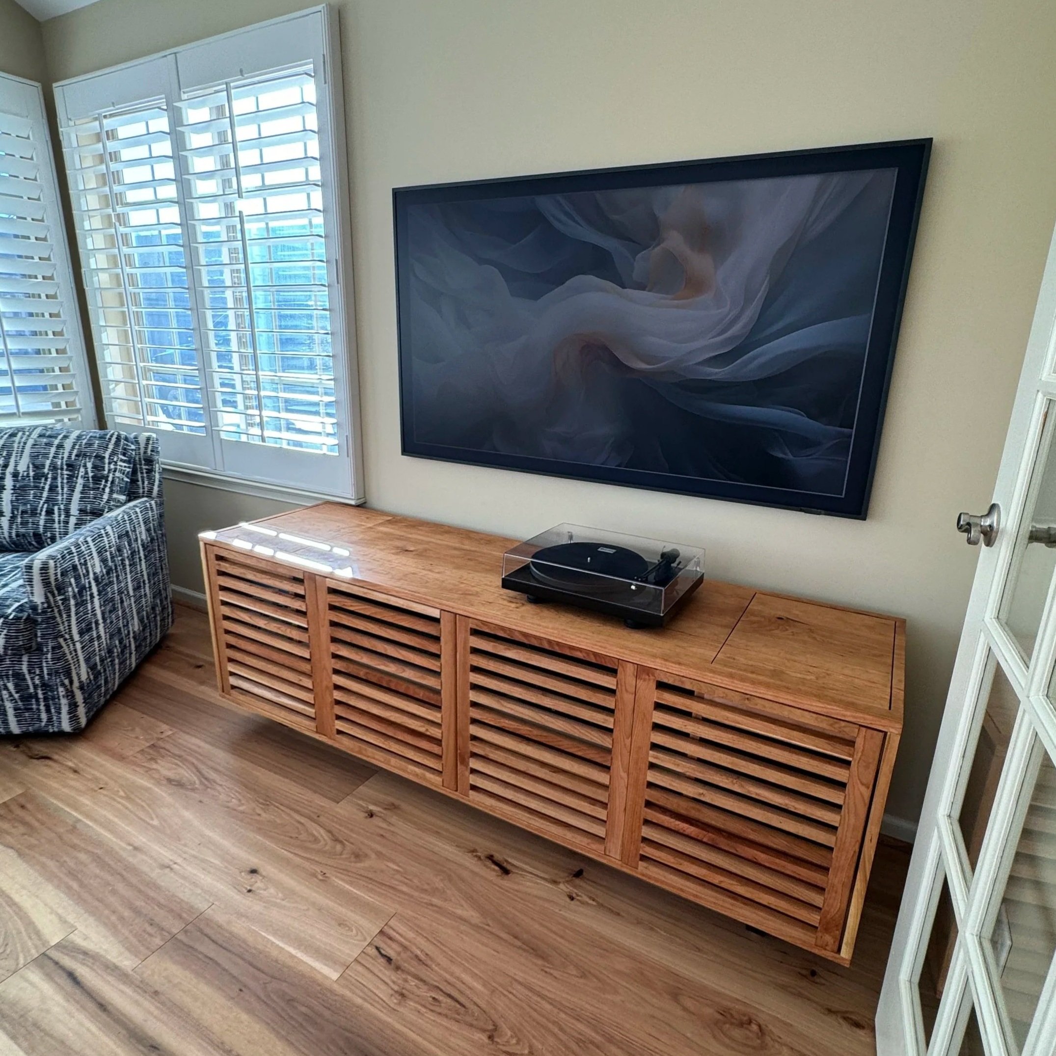 A living room corner featuring a wooden slatted TV stand with a flat-screen TV and a turntable on top, next to a window with white plantation shutters and a patterned armchair.