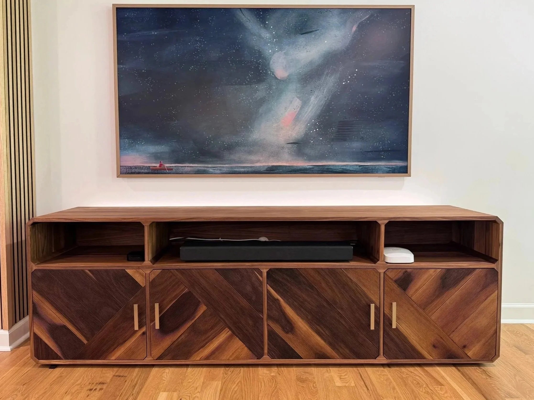A colorful abstract painting with a night sky, stars, and a small red boat on water, framed and hung on a white wall above a wooden cabinet with geometric patterns.
