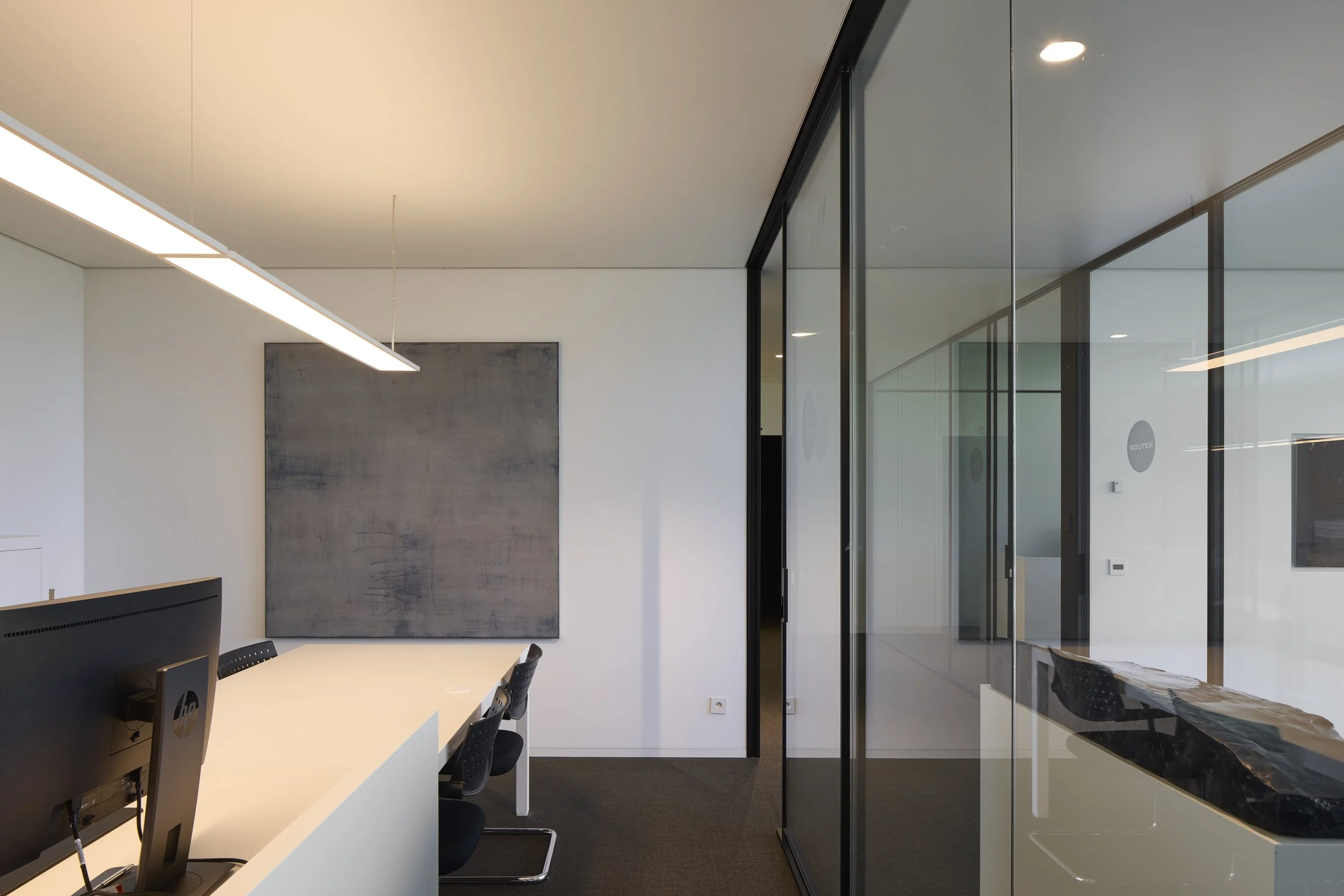 Modern office interior with a white table, black chairs, a gray abstract wall art, and a glass wall with black frames, illuminated by ceiling lights.