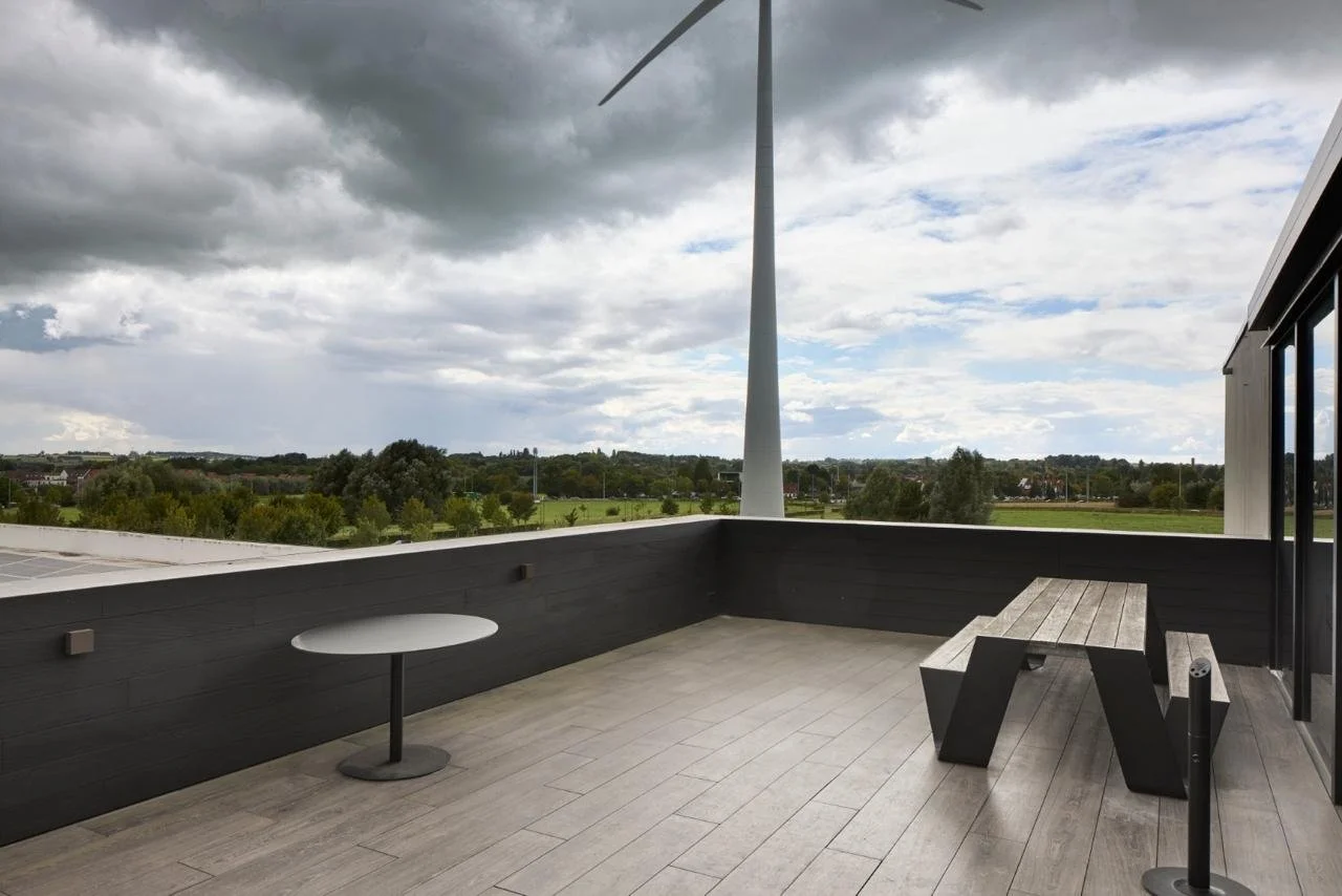 Empty balcony with a modern black and wooden bench, small round table, black railing, wind turbine in the background, and cloudy sky.
