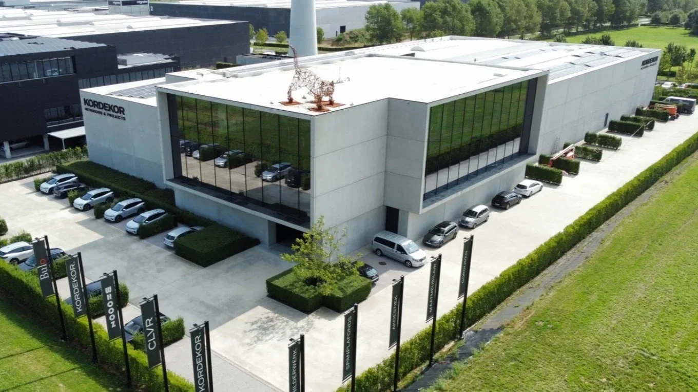 A modern commercial building with reflective glass windows, surrounded by a parking lot with several cars, green bushes, and a landscaped exterior with signage displaying 'KORDEKOR'.