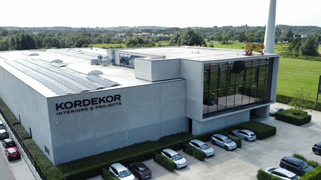Aerial view of a modern commercial building labeled 'KORDEKOR Interiors & Projects' with a parking lot and solar panels on the roof, surrounded by greenery.