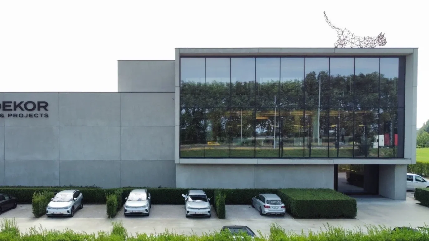 Modern building with large glass window reflecting trees, parked cars in front, and a wire sculpture of a cat on the roof.