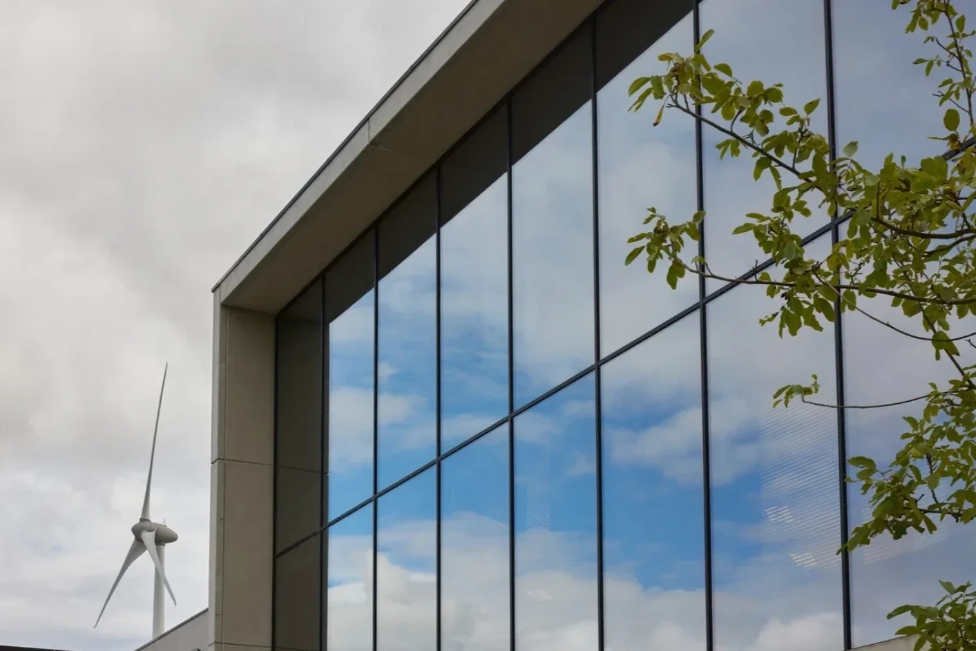 Modern building with large glass windows reflecting the sky and clouds, with a wind turbine in the background and some tree branches in the foreground.