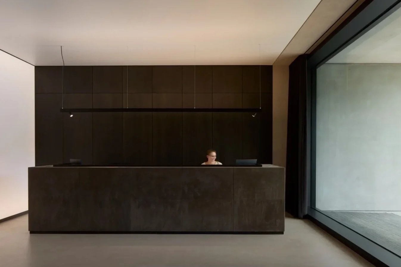 Minimalist reception area with a dark-colored front desk and a person sitting behind it, set against a black wall, with a large glass window to the right.
