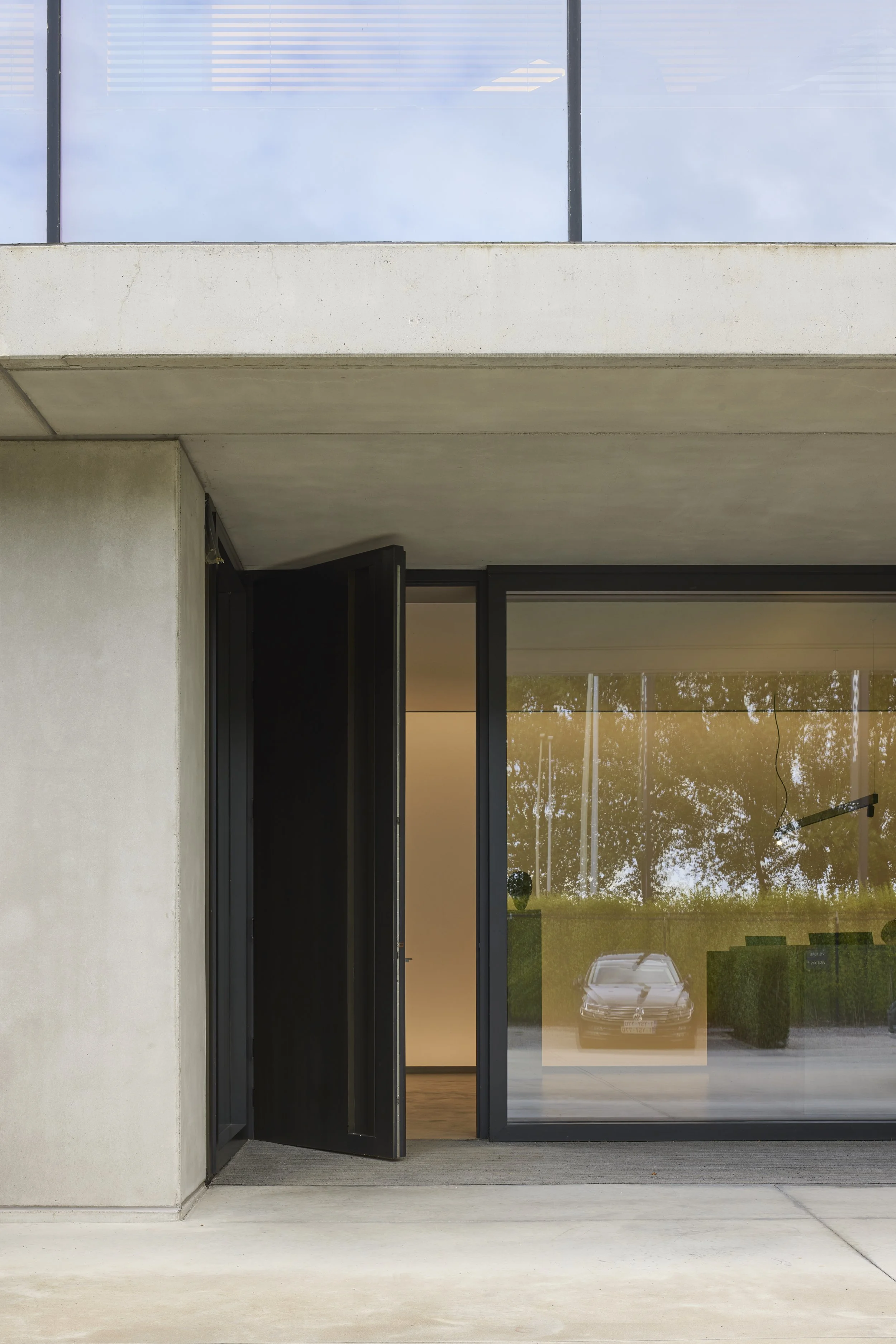 Modern building exterior featuring a glass door with a black frame, adjacent to a concrete wall, and a large window showing reflections of trees and parked cars outside.