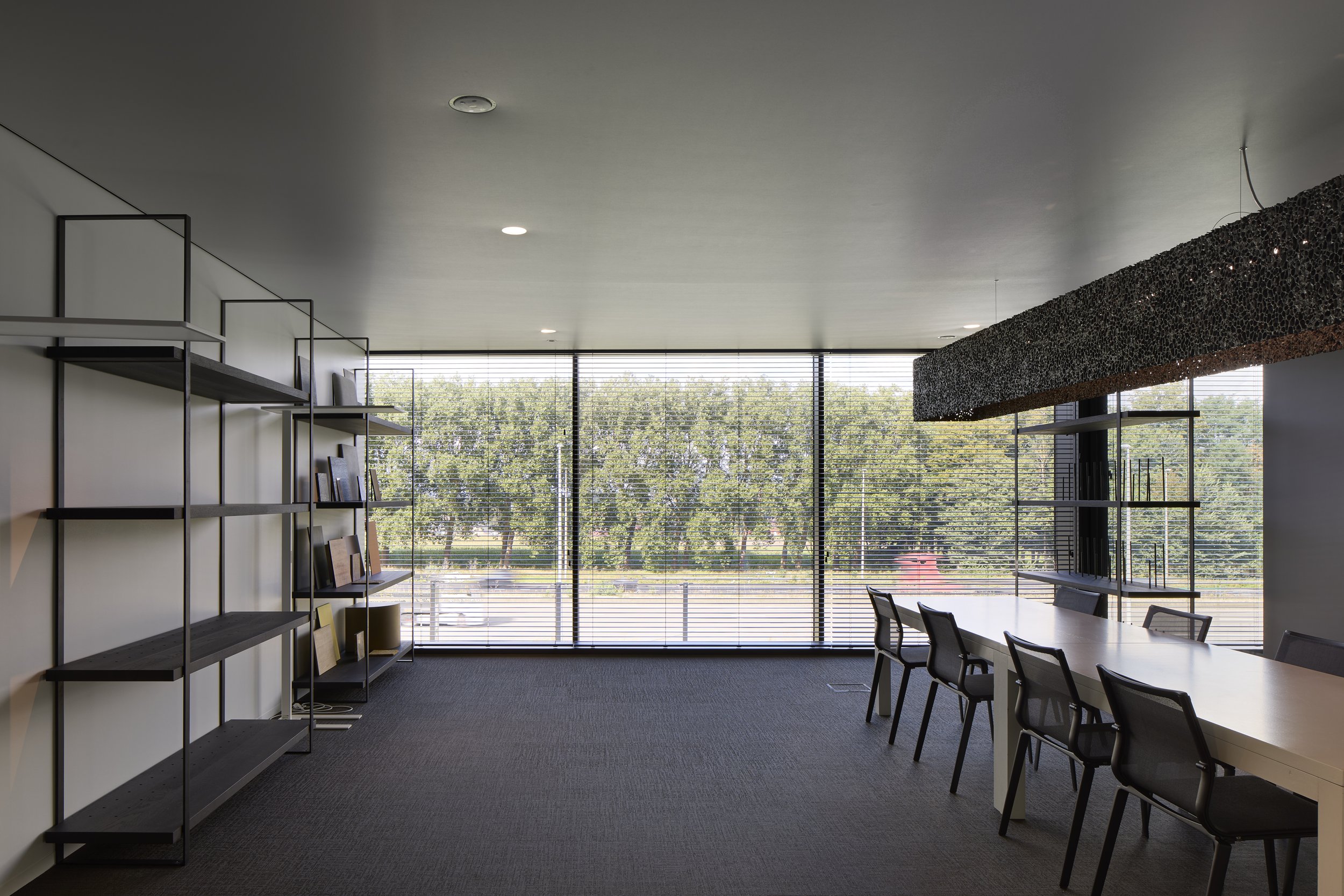 Interior of a modern conference room with large windows, a long white table surrounded by black chairs, and dark trim shelves with books and decor. A textured black light fixture hangs from the ceiling.