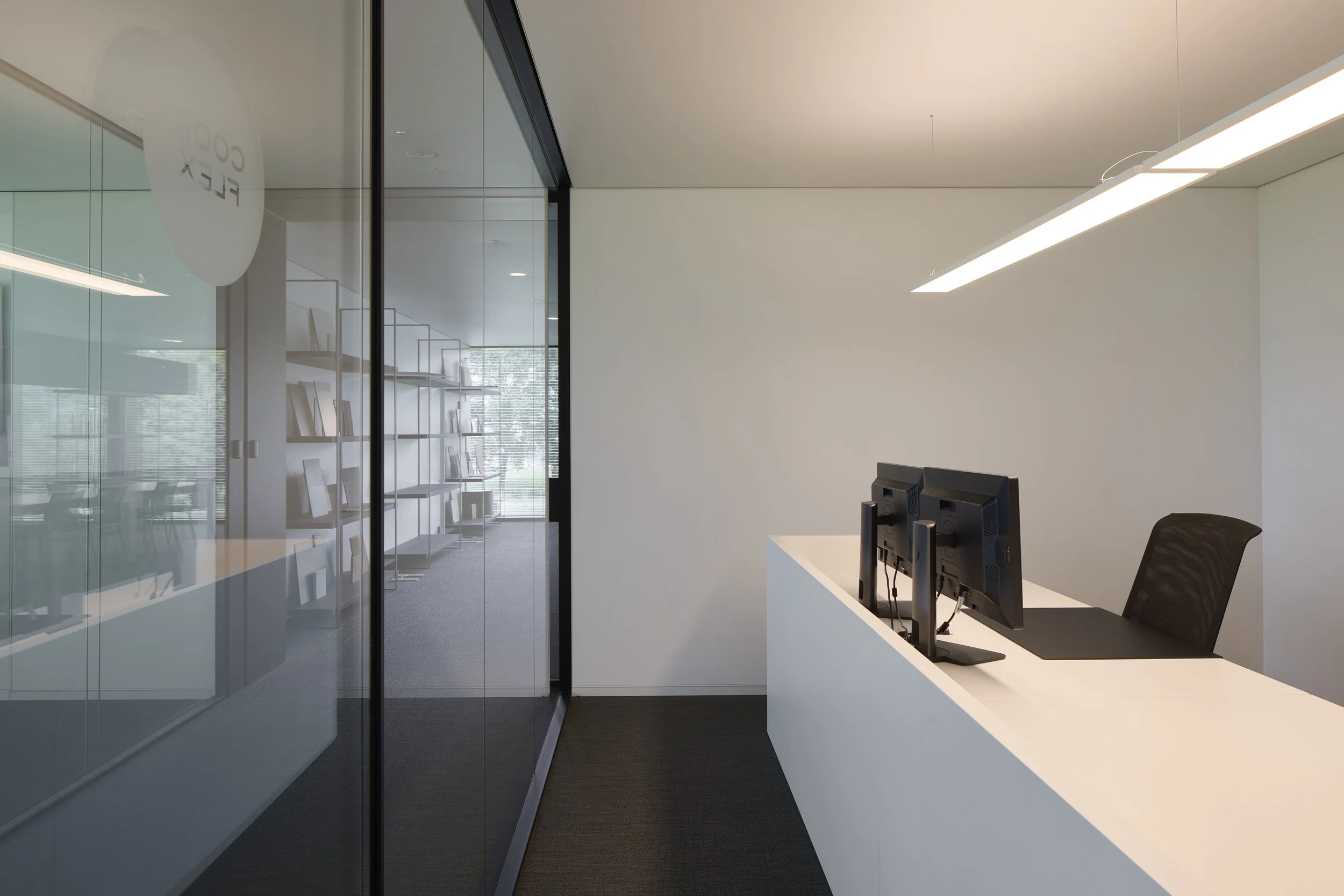 Modern office space with a white desk, two computer monitors, a black office chair, and a glass wall with shelves and books inside.