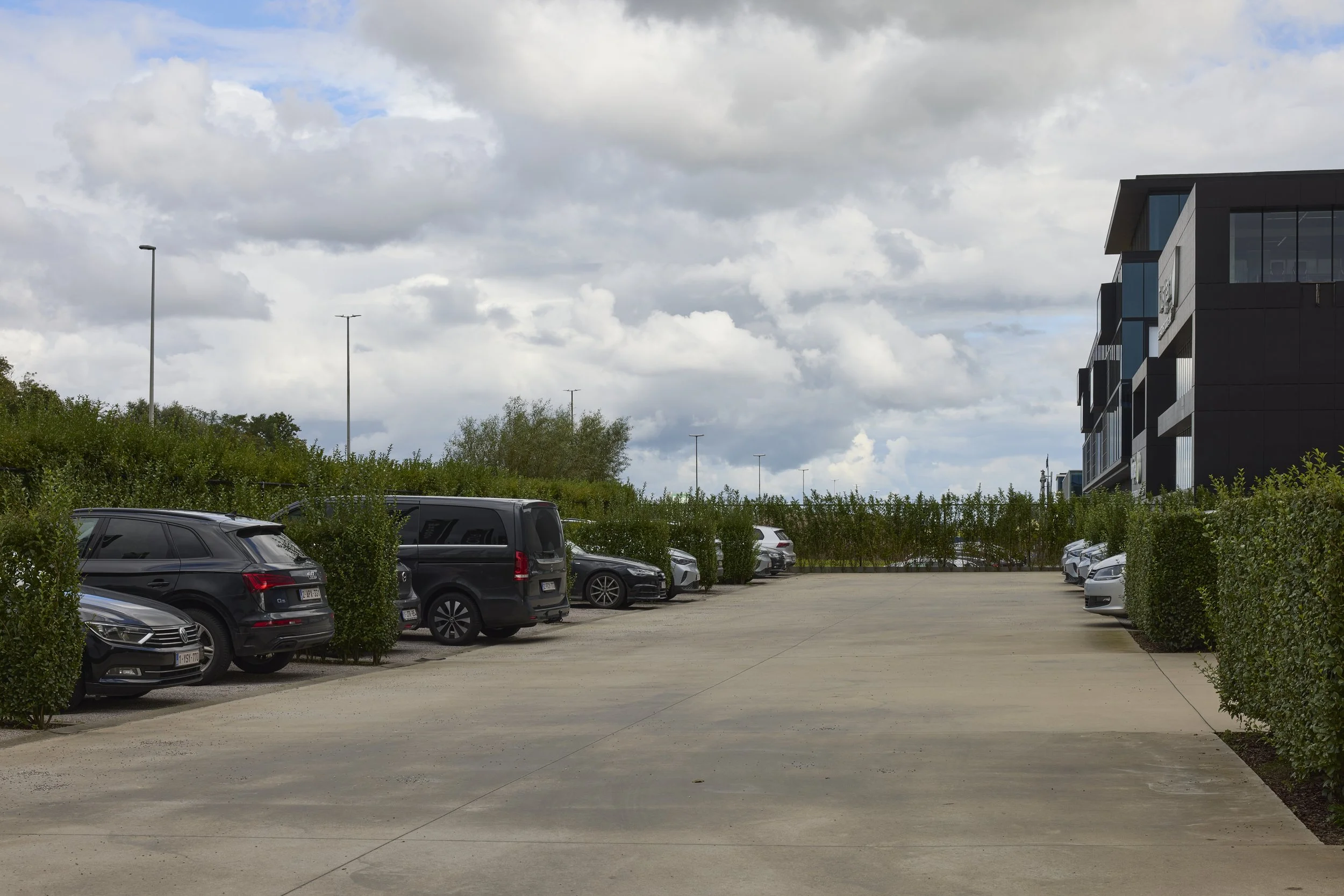 A parking lot with several parked cars, surrounded by green bushes, with a modern black building on the right and a cloudy sky overhead.