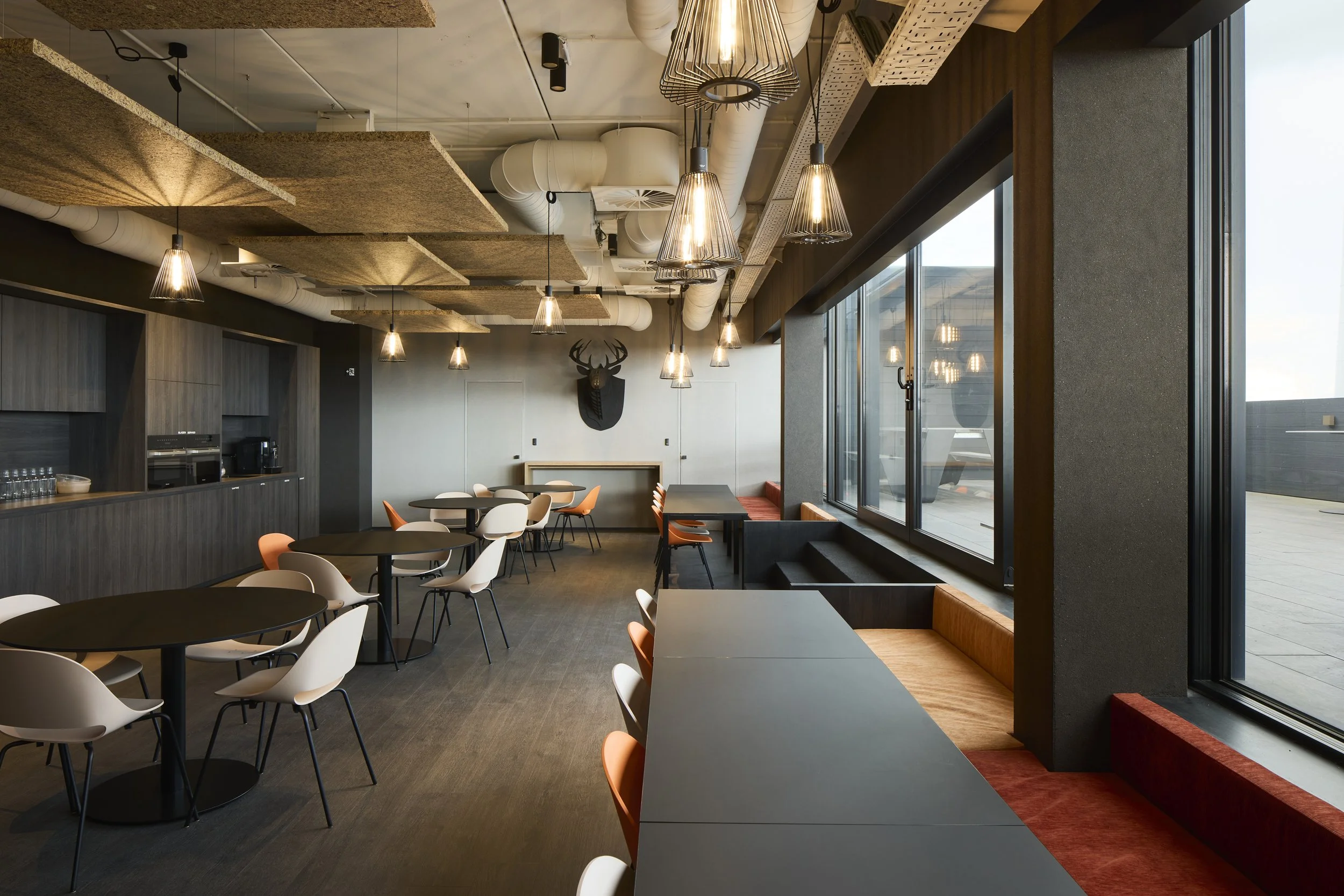 Modern dining area with black tables and white and orange chairs, large windows, pendant lights, and a wall decoration of a deer head.