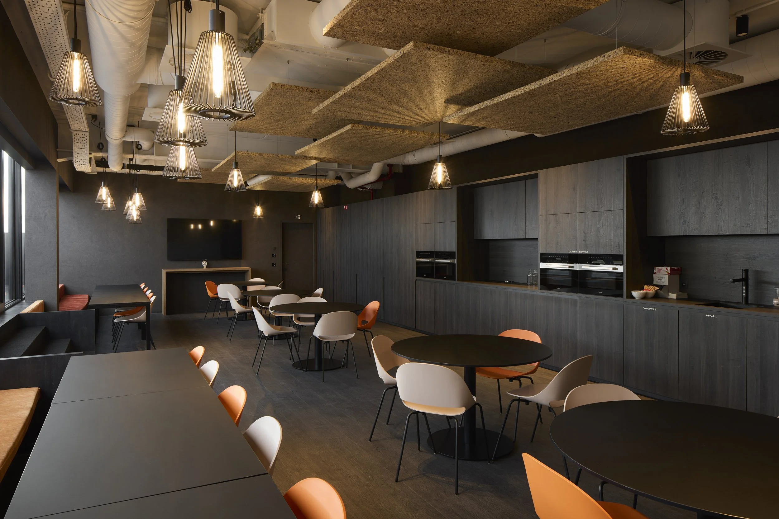 Modern indoor dining or conference room with dark round tables, white and orange chairs, wood-paneled cabinets, wall-mounted TV, and unique ceiling acoustic panels with hanging lights.