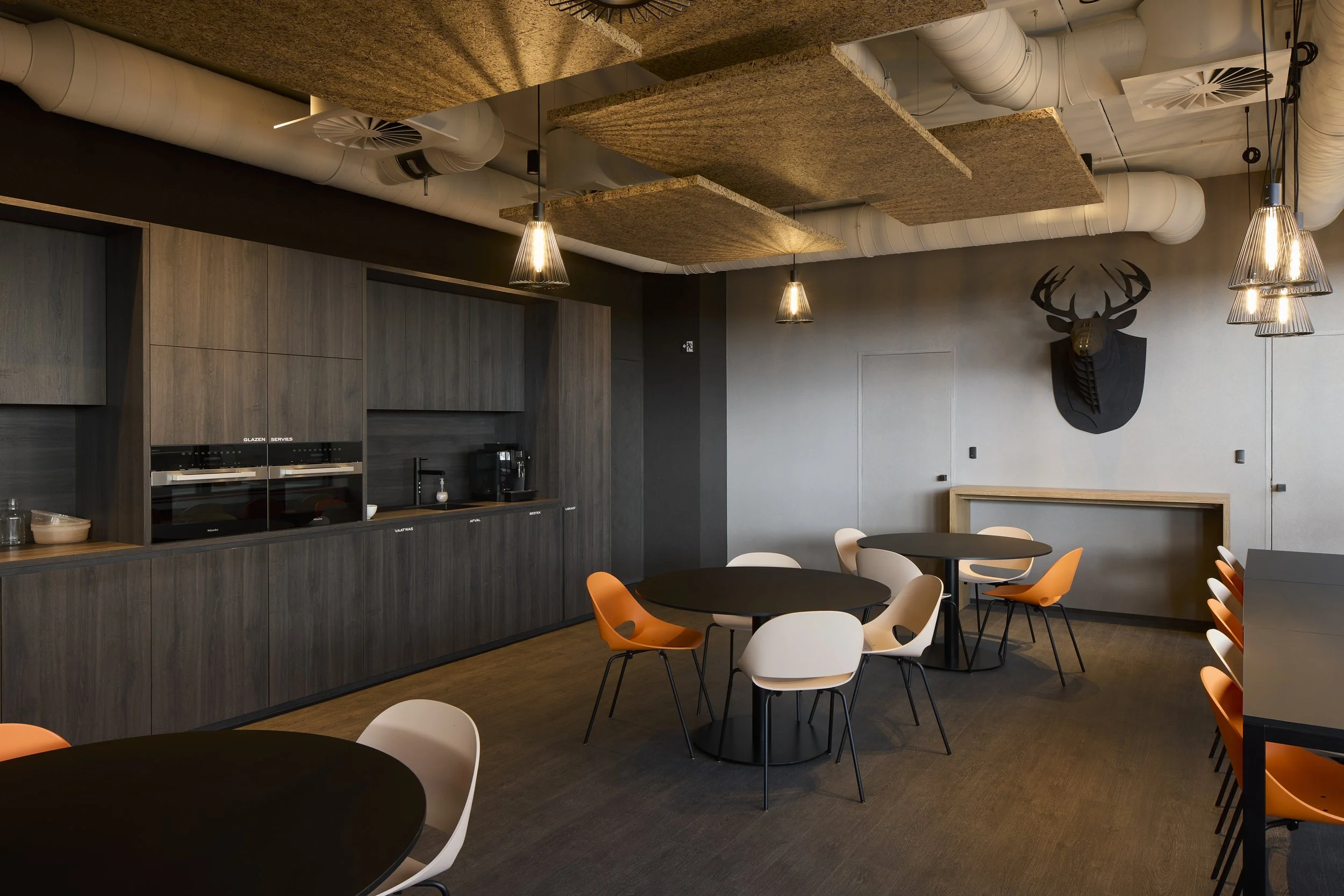 Modern dining area with black round tables and white and orange chairs, wood-paneled cabinet with built-in appliances, decorative deer head wall art, ceiling with acoustic panels and exposed ductwork, and pendant lighting.