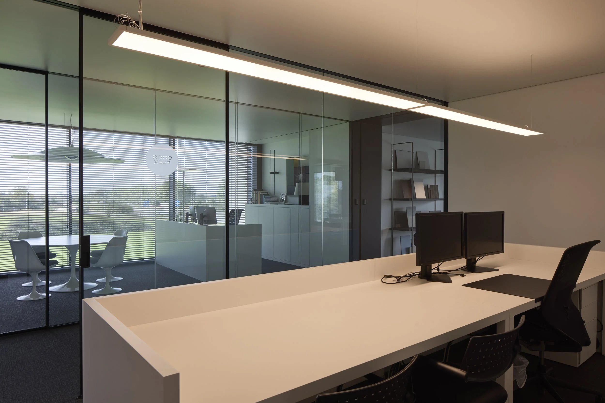 Modern office space with a white desk featuring two computer monitors, surrounded by black chairs, with a large glass wall revealing a meeting area with a round table and white chairs. The background shows an office shelf with books and artwork, larg