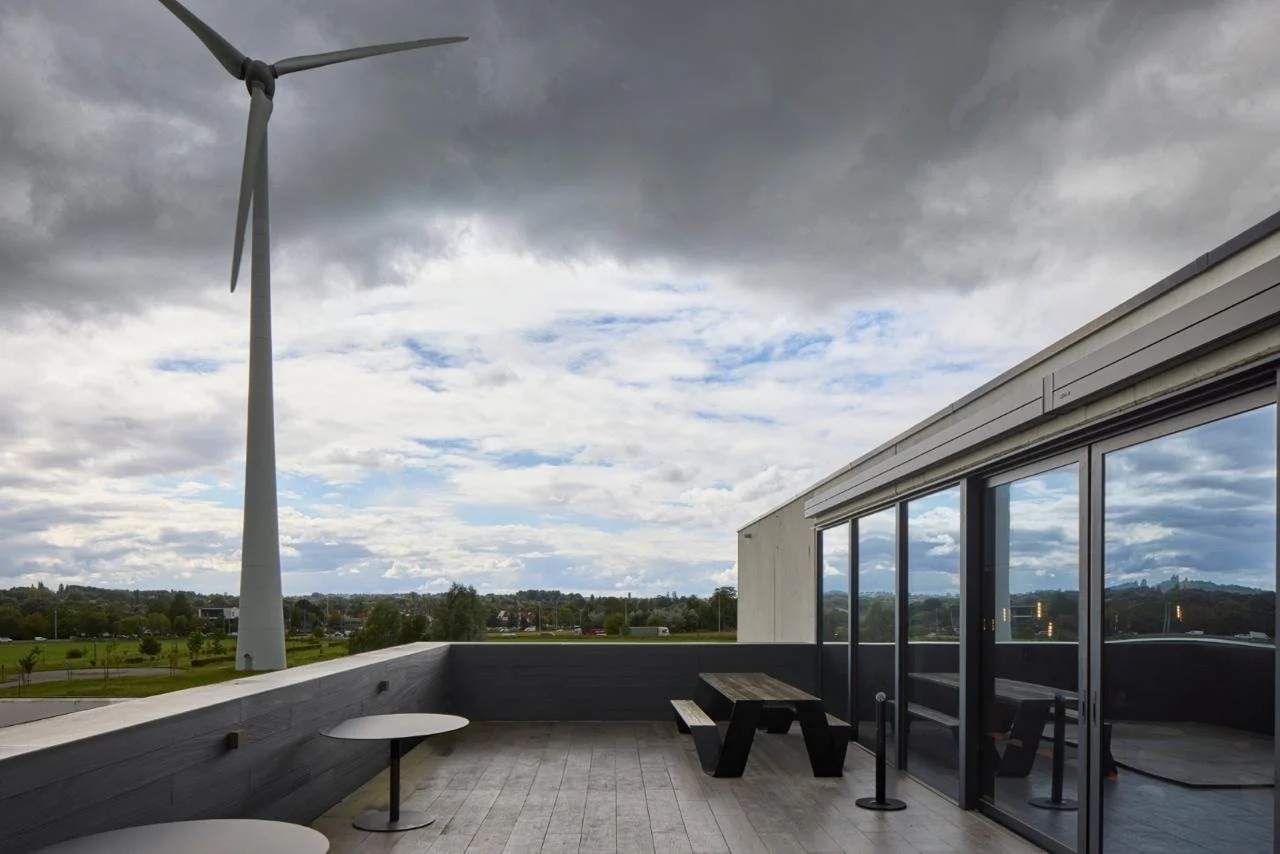 View from a balcony with wind turbines and cloudy sky, railing, glass doors, benches, and tables.