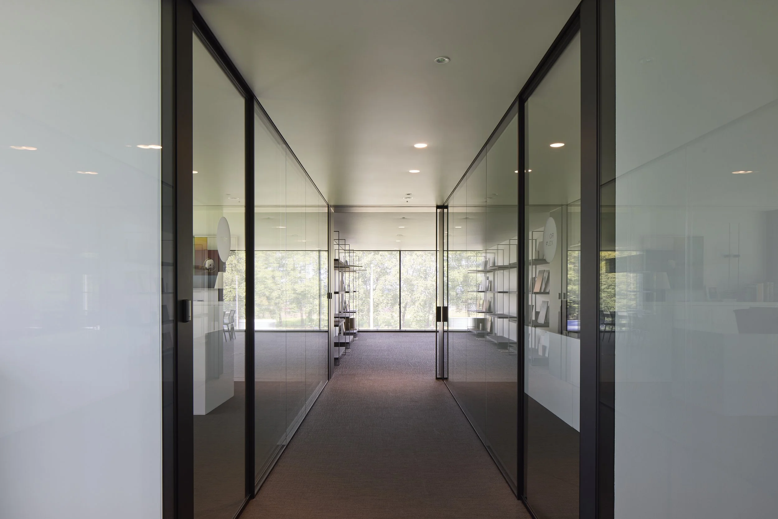 View through glass walls of a modern office interior with shelves and large windows showing greenery outside.