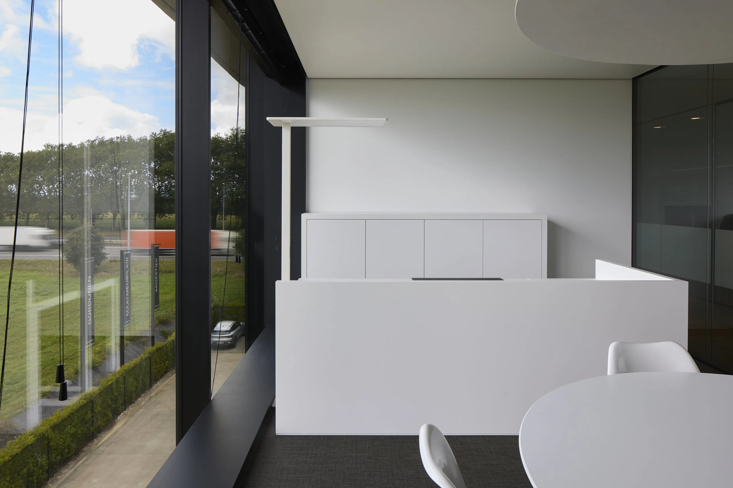 Interior of a modern building with a white table and white chairs, large floor-to-ceiling glass windows showing trees and a road outside.
