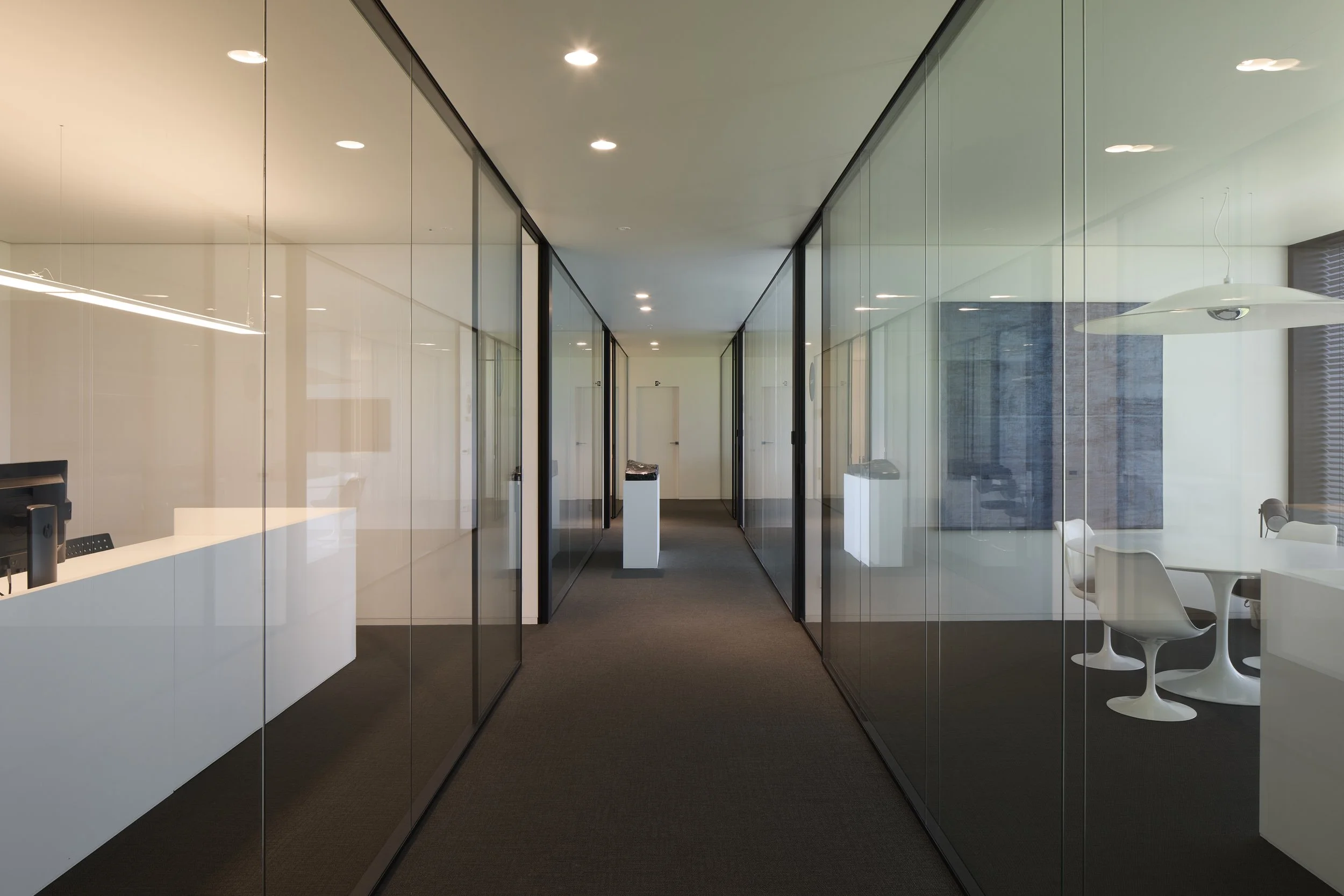 Modern office corridor with glass walls and doors, white reception desk on the left, and seating area with white chairs on the right. Ceiling lights illuminate the space.